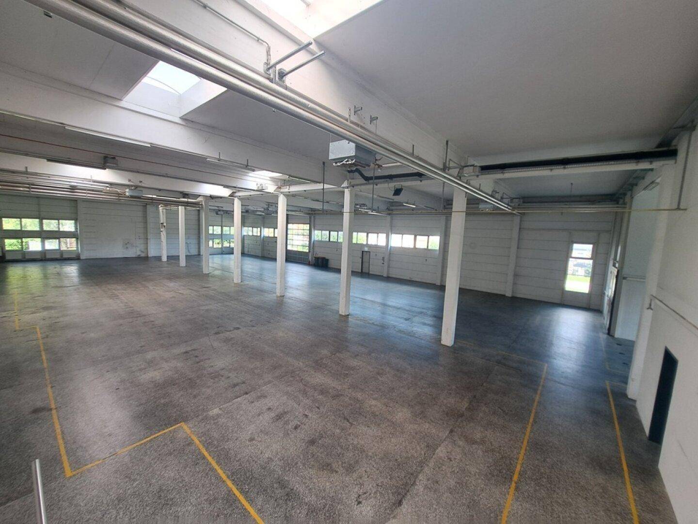 Geräumige Lagerhalle mit Betonboden, Stützpfeilern, Oberlichtern und Rolltoren für viel Tageslicht.