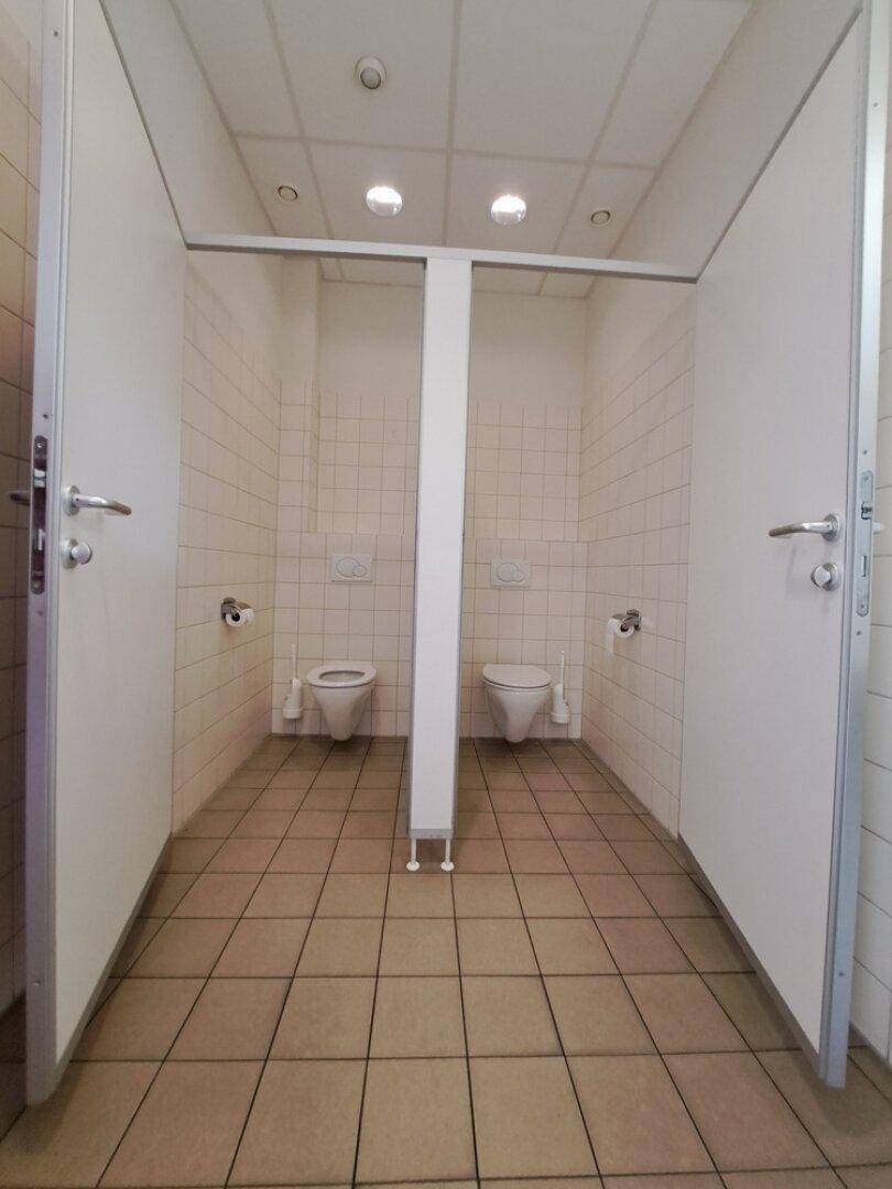 Öffentliche Toilettenanlage mit zwei Kabinen, gefliesten Wänden und Boden, sauber und funktional.