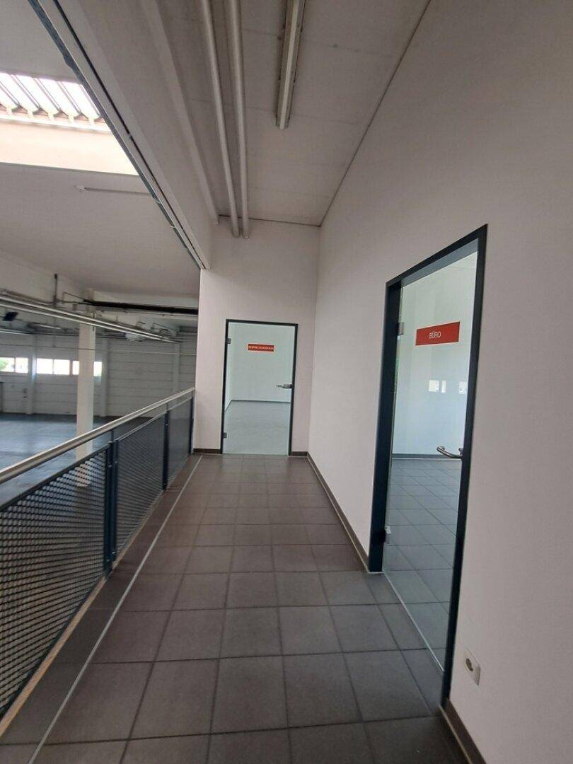 Flur mit dunklen Fliesen, weißen Wänden und einem Geländer mit Blick auf die Lagerhalle.