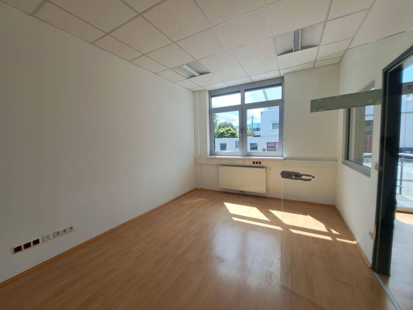 Leeres Büro mit hellem Laminatboden, weißen Wänden und großen Fenstern für viel Tageslicht.