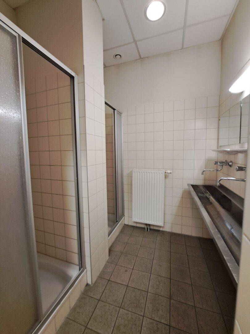 Öffentlicher Duschraum mit mehreren Duschkabinen, gefliesten Wänden und Boden sowie einem langen Waschbecken.