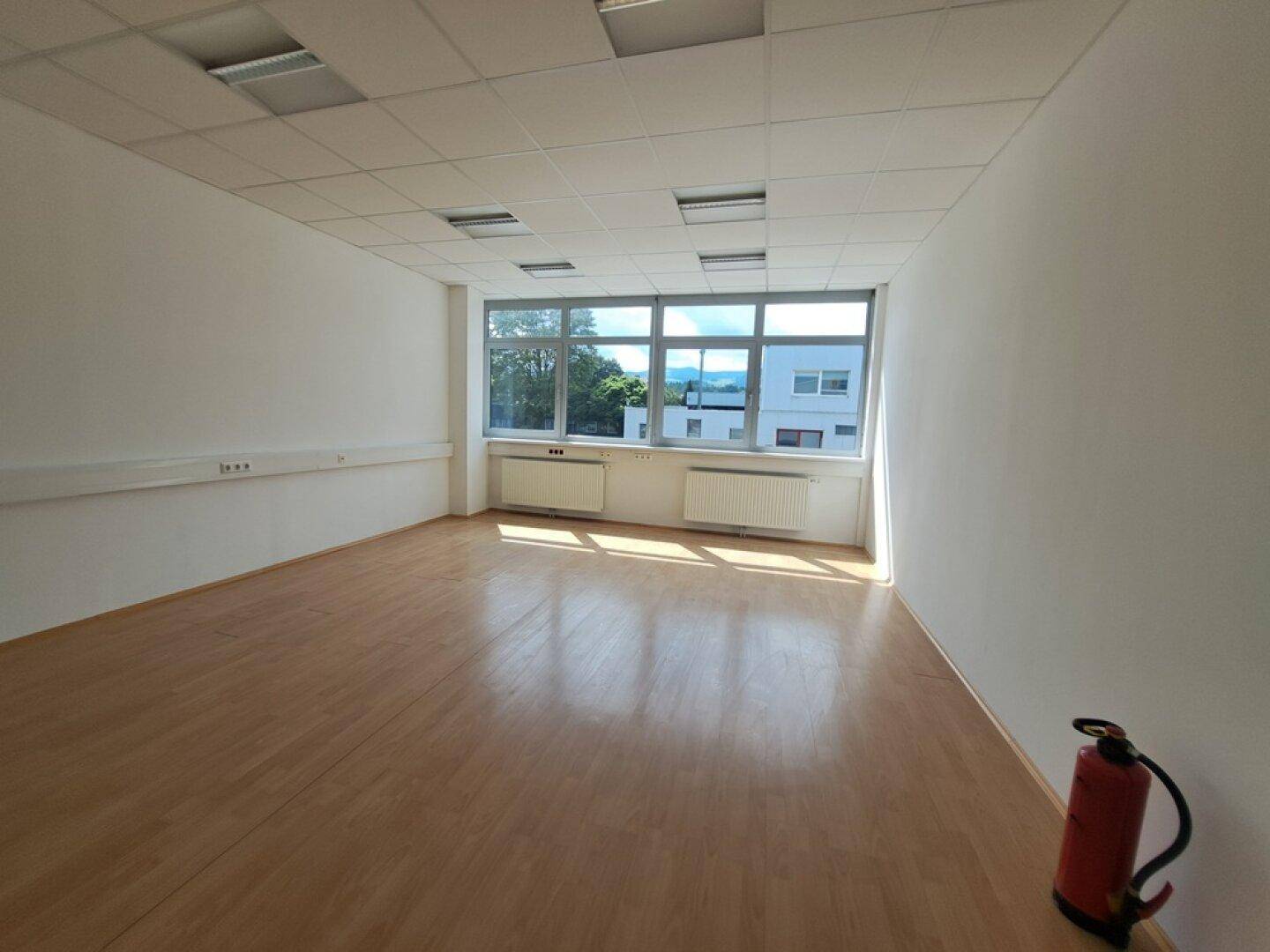 Geräumiges Büro mit Laminatboden, großen Fenstern und einem Feuerlöscher, gut beleuchtet.