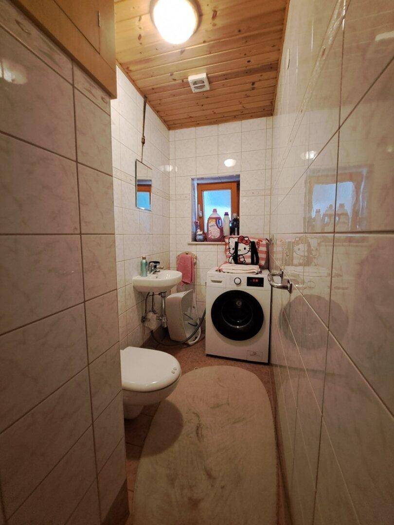 Kleines WC mit Waschbecken, Toilette, Waschmaschine und gefliesten Wänden sowie Holzdecke.