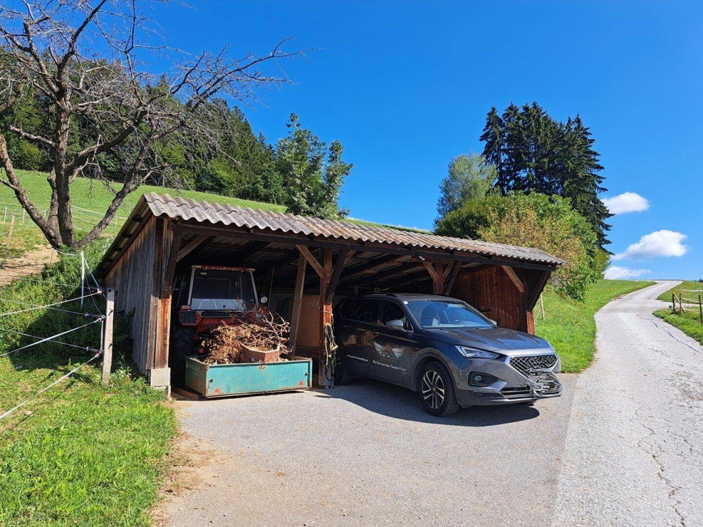 Offener Holzunterstand dient als Carport für ein Auto und landwirtschaftliche Geräte.