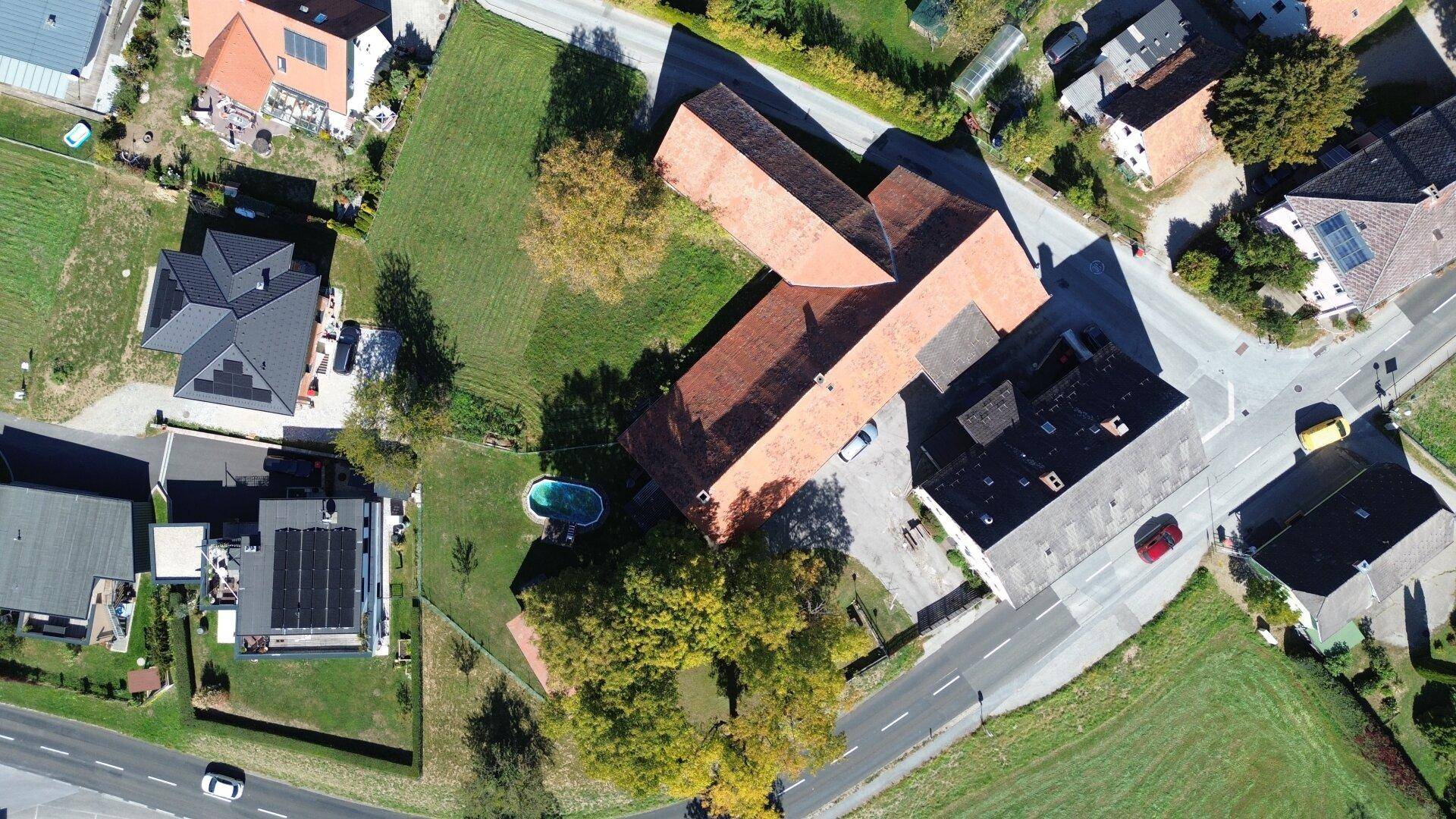 Draufsicht auf das Anwesen mit Hauptgebäude, rotem Scheunendach, kleinem Pool und angrenzenden Straßen und Häusern.