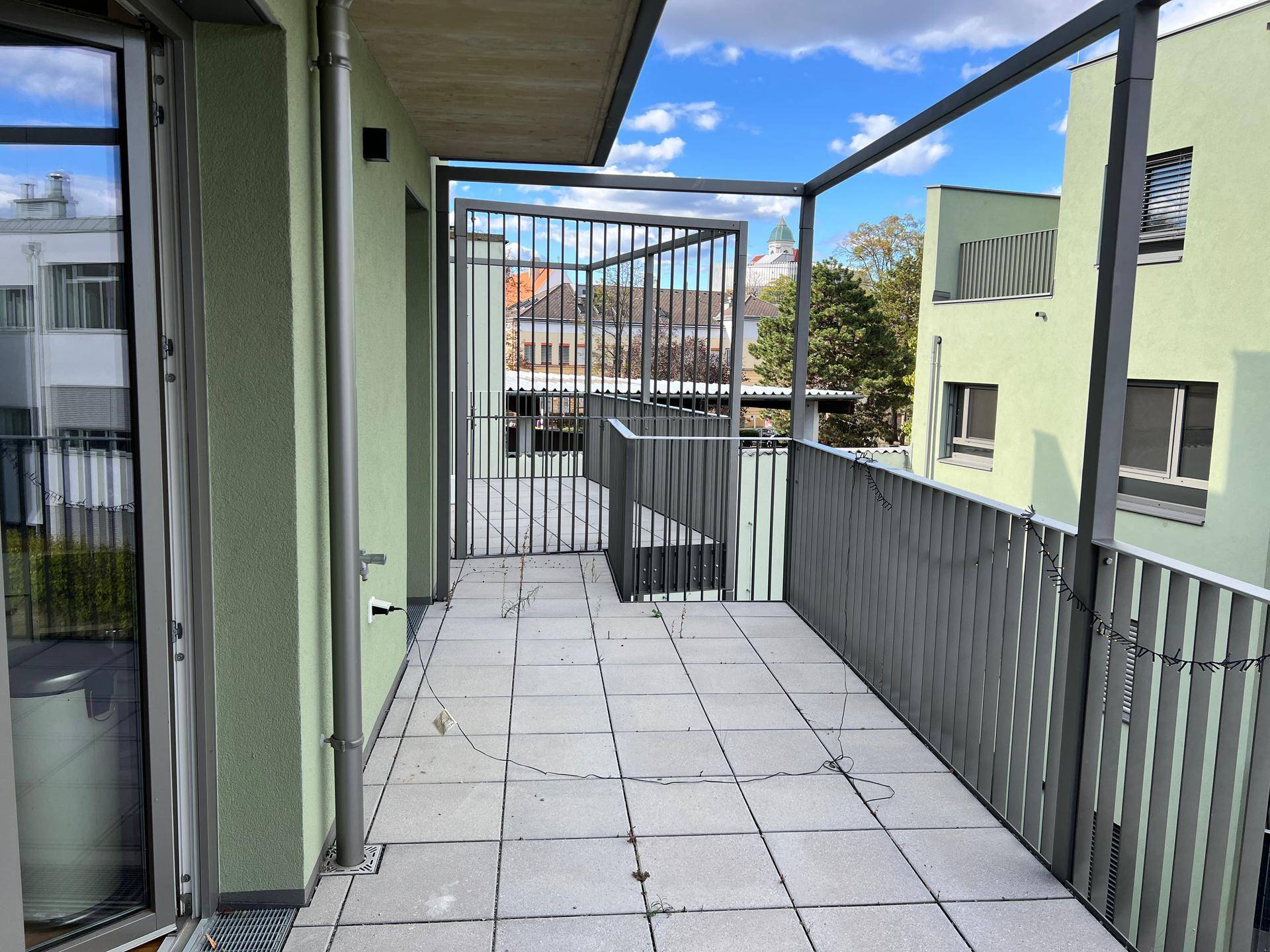 Breiter Balkon mit Fliesenboden und Metallgeländer, umgeben von modernen Gebäuden.