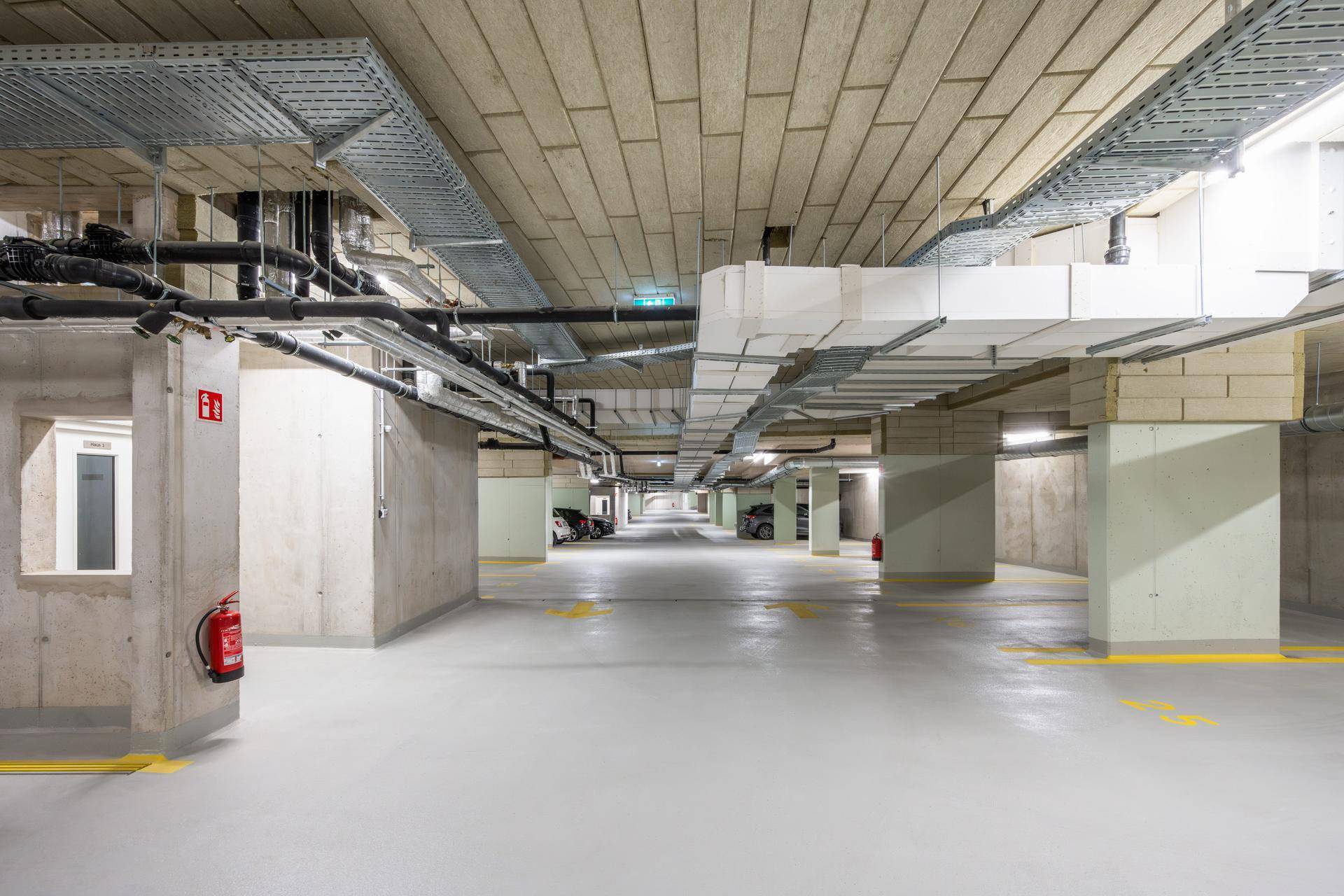 Geräumige Tiefgarage mit Betonwänden, sichtbaren Leitungen und klarer Beschilderung auf dem Boden.
