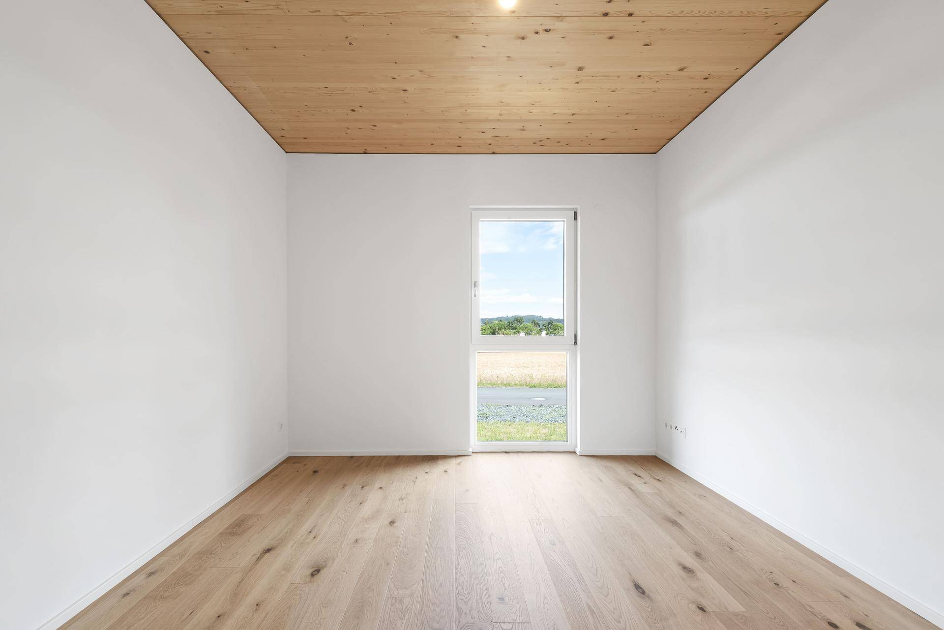 Ruhiges Schlafzimmer mit Holzboden, Holzbalkendecke und einem Fenster mit Blick ins Grüne.