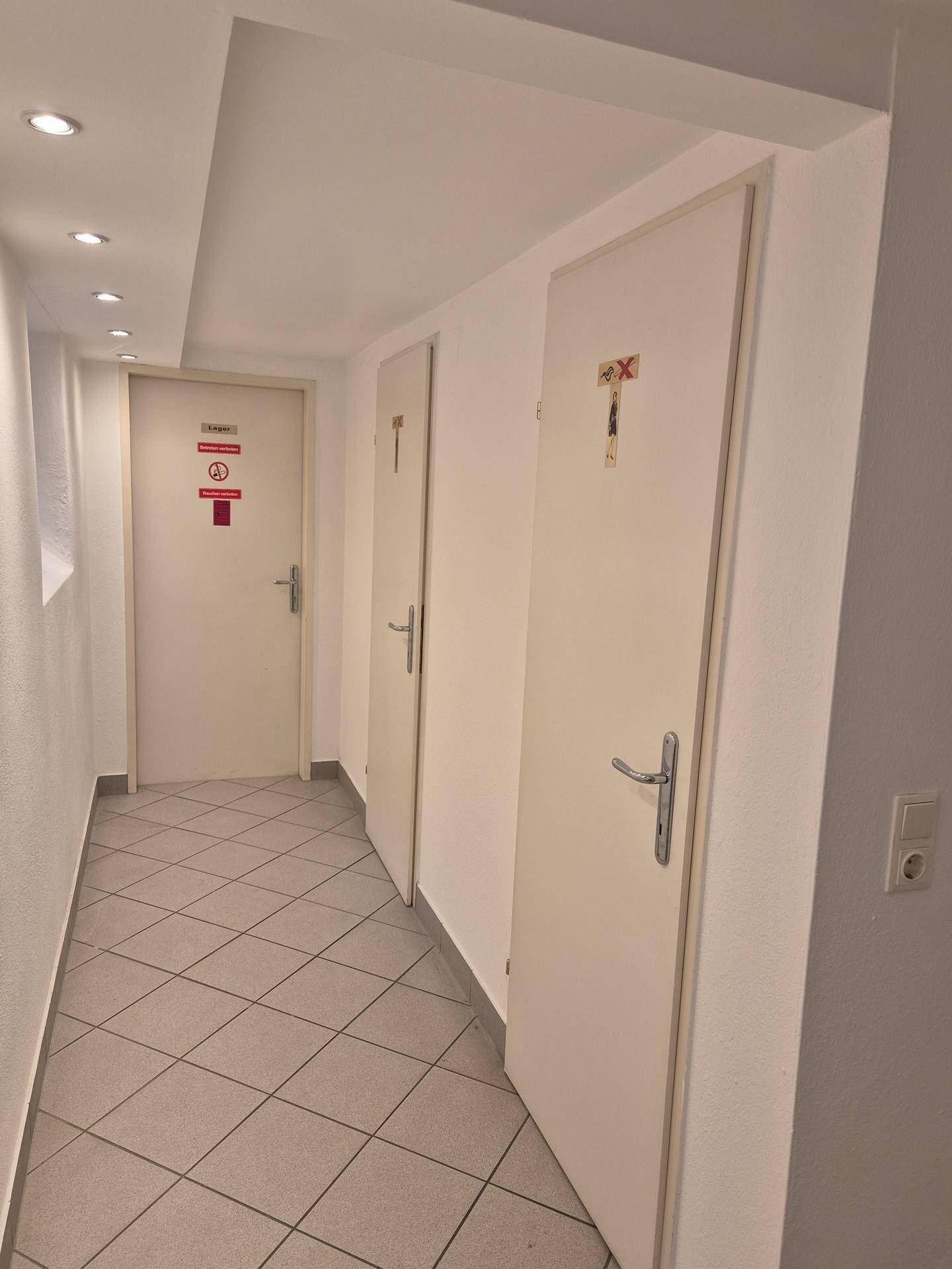Flur mit Fliesenboden und mehreren Türen, darunter Toiletten, beleuchtet durch Deckenspots.