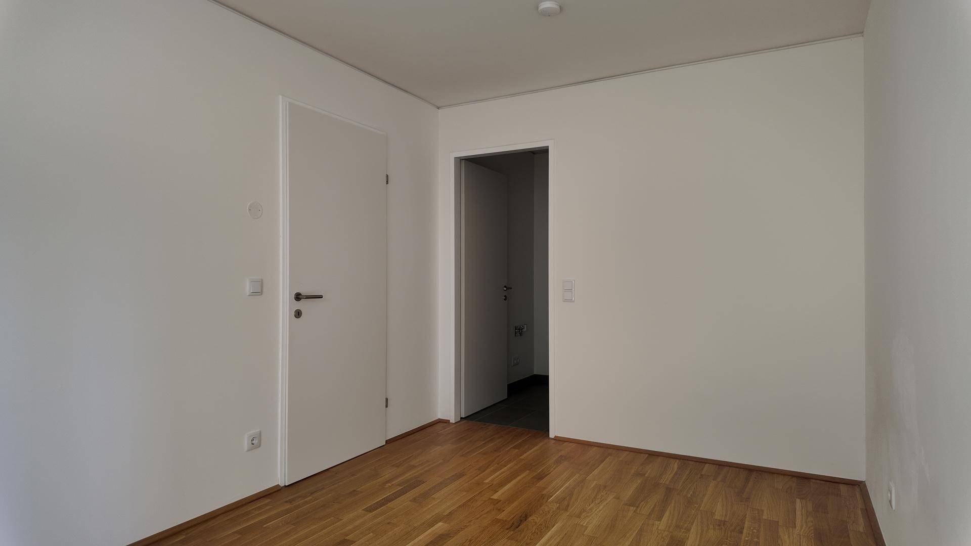 Heller Raum mit Holzboden und zwei weißen Türen, eine davon führt in einen angrenzenden Bereich.