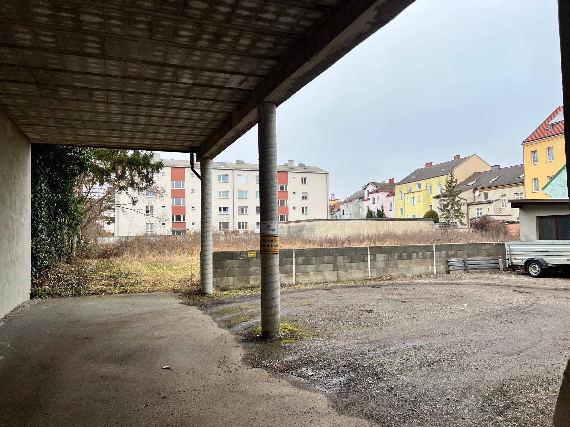 Überdachter Bereich mit Betonpfeilern und Blick auf ein unbebautes Grundstück und Wohngebäude.