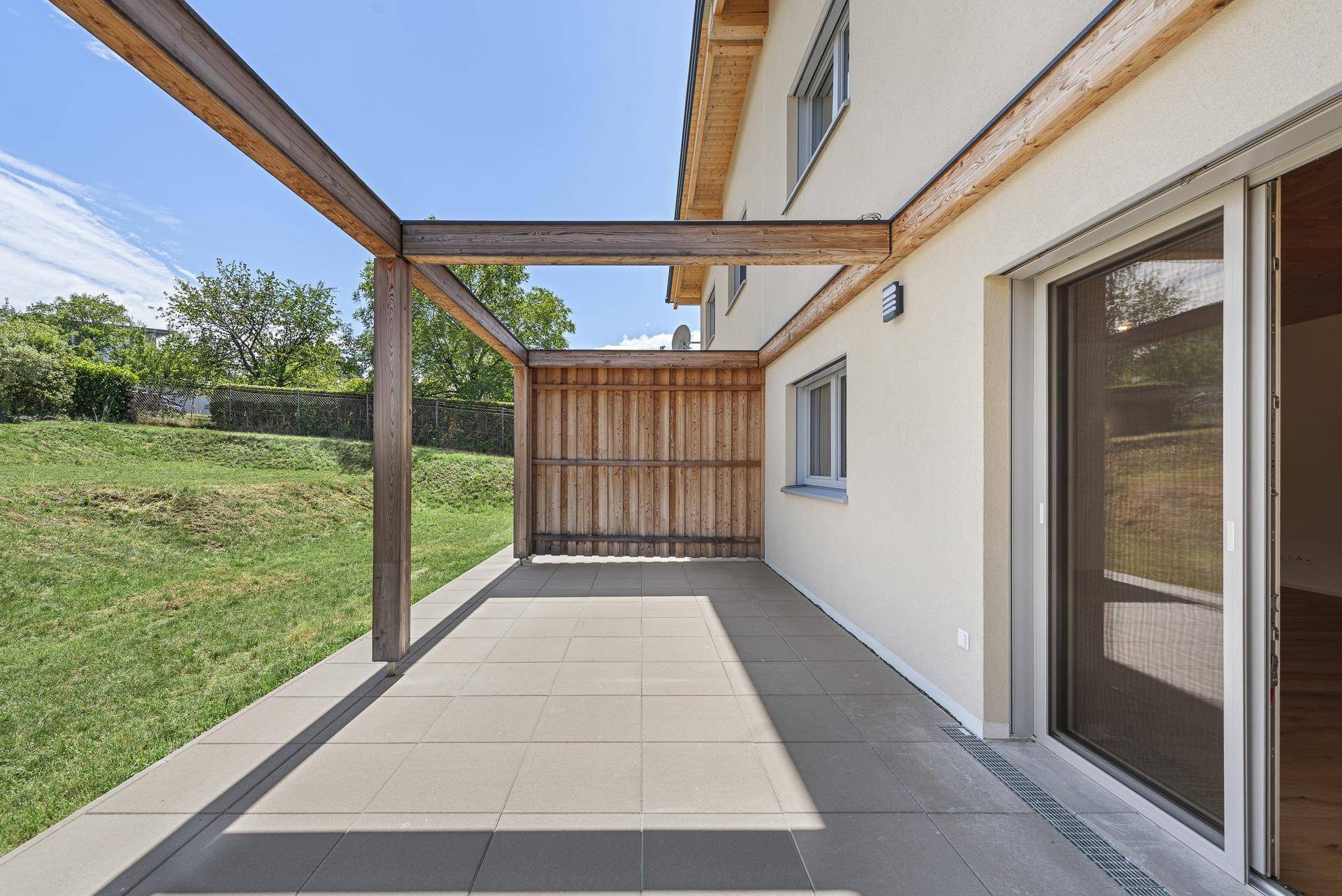 Sonnige Terrasse mit Fliesenboden, Holzpergola und angrenzender Grünfläche.