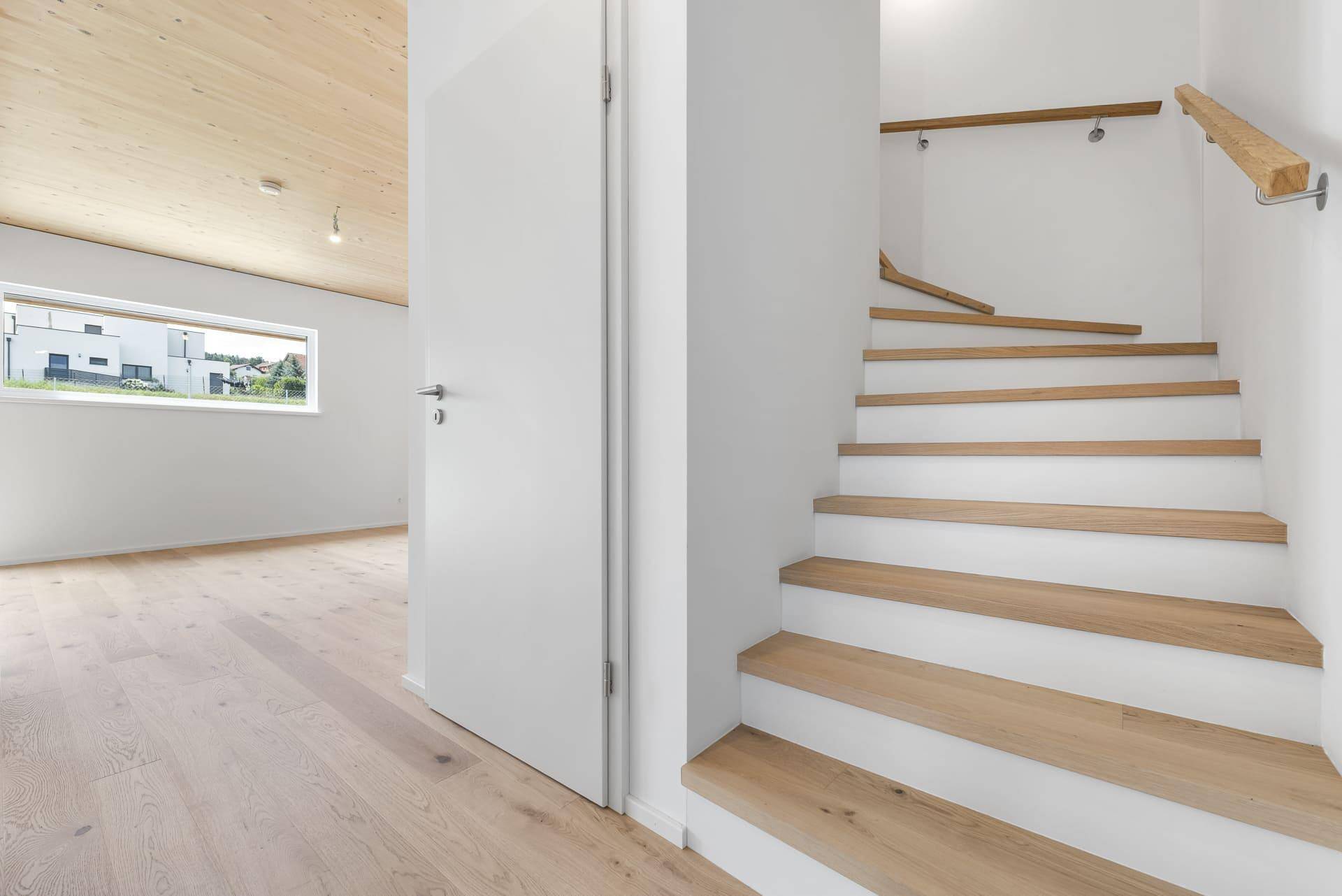 Moderne Holztreppe mit weißem Geländer und Blick aus dem Fenster in die grüne Umgebung.