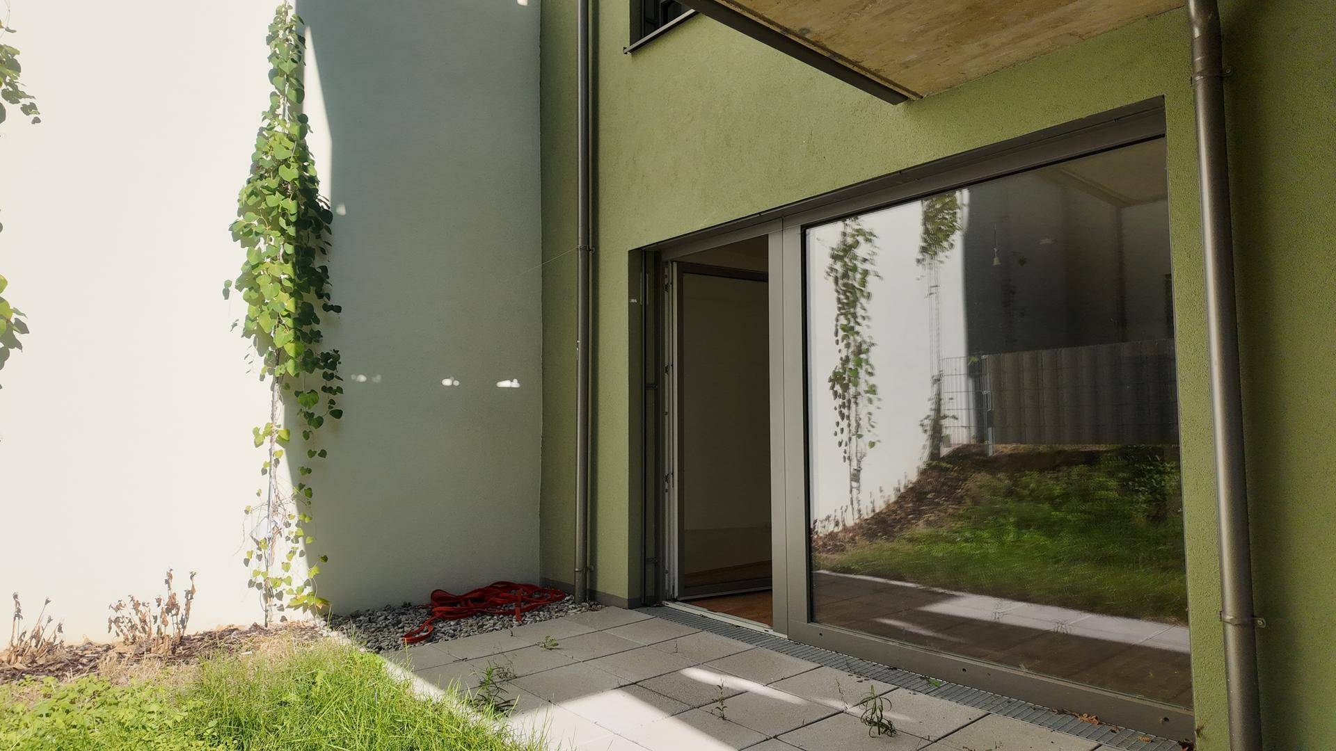 Außenbereich mit gepflasterter Terrasse, bewachsener Wand und großem Fenster zum Innenraum.