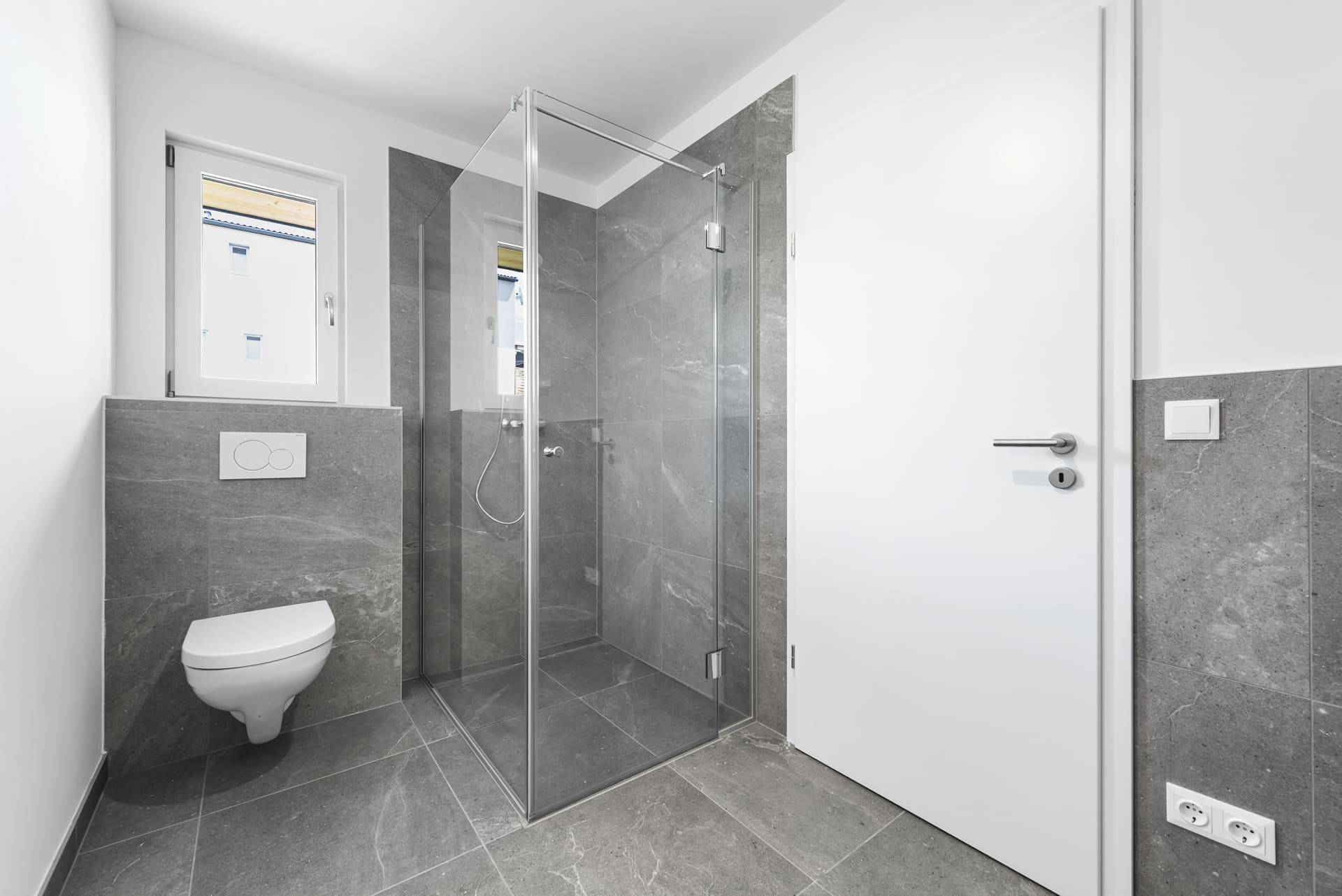 Modernes Badezimmer mit grauen Fliesen, wandhängendem WC und einer geräumigen Glasdusche.