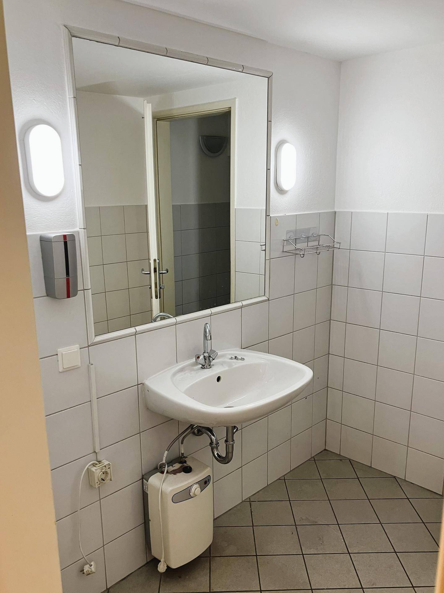 Einfaches Badezimmer mit Waschbecken, Spiegel und weißen Fliesen an Wänden und Boden.