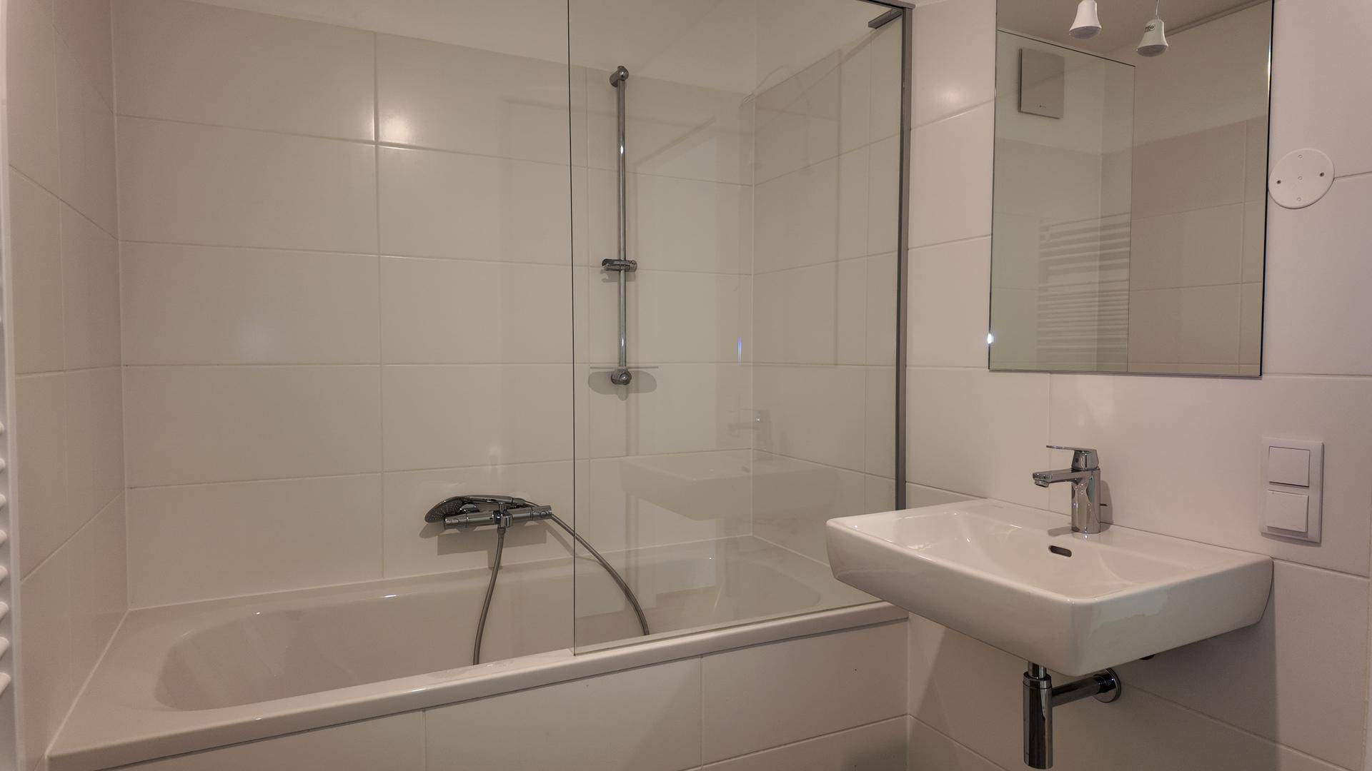 Helles Badezimmer mit Badewanne, Duschvorrichtung, Waschbecken und Spiegel über dem Waschtisch.