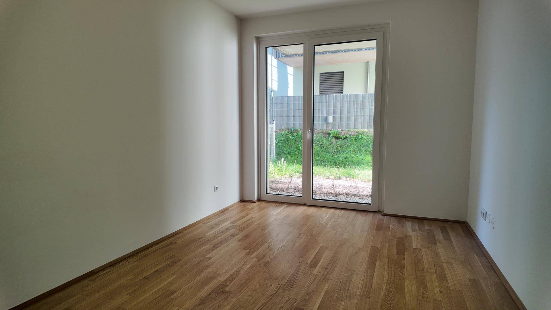 Leeres Schlafzimmer mit Holzboden und direktem Zugang zu einem kleinen Grünbereich durch eine Glastür.