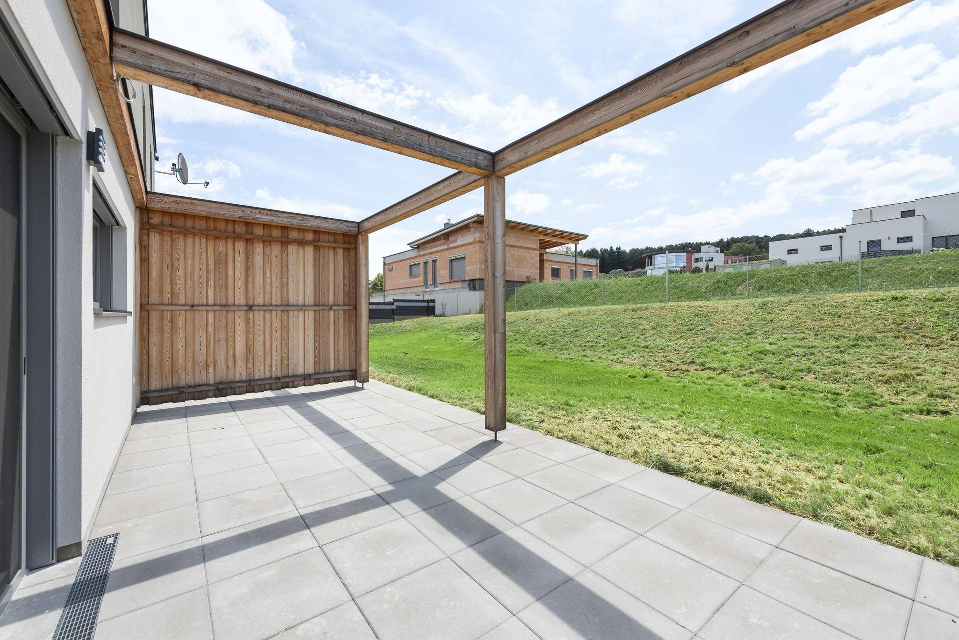 Geräumige Terrasse mit Betonplatten und einer Holzpergola, umgeben von Grünflächen.