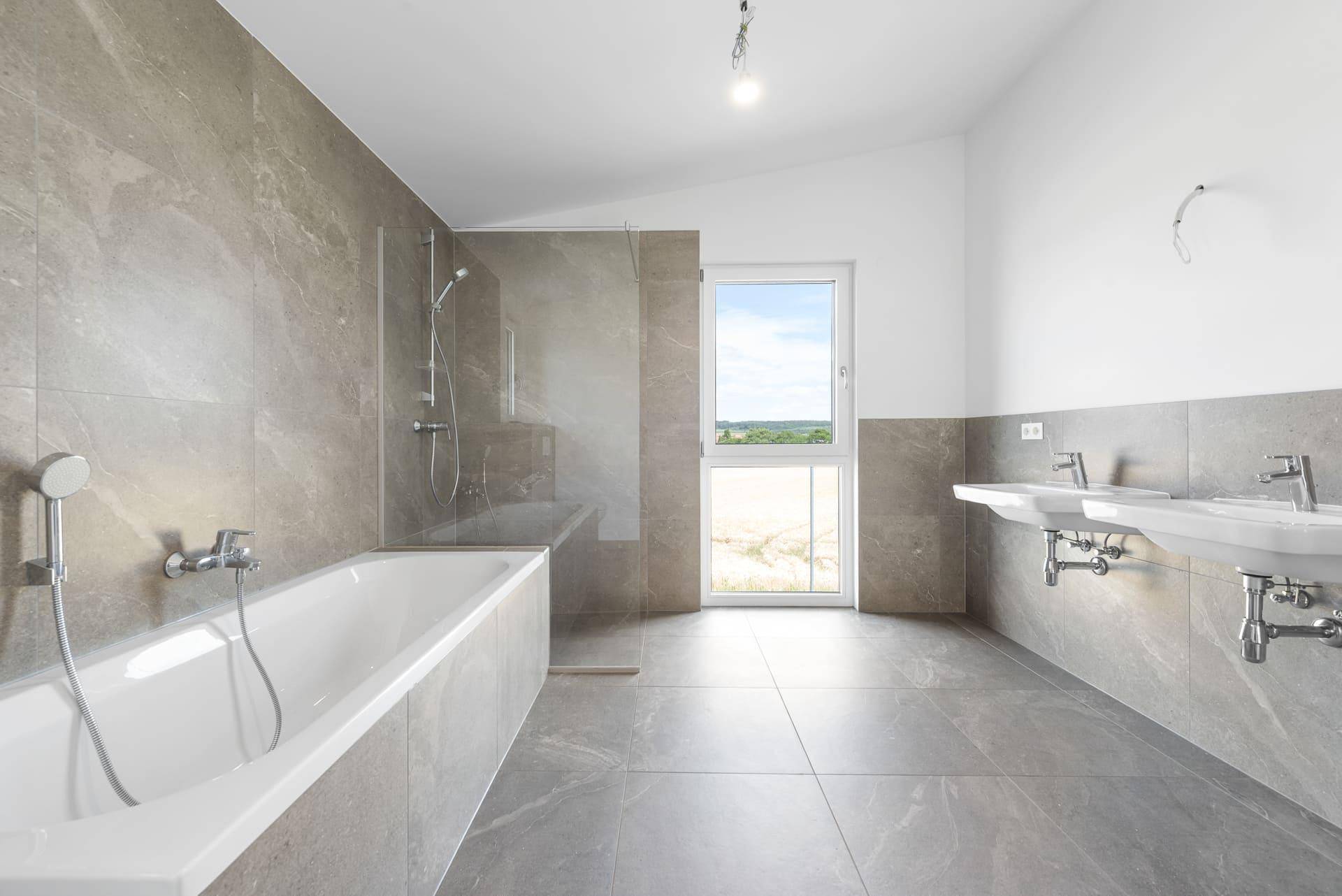 Großes Badezimmer mit grauen Fliesen, Badewanne, Dusche und Doppelwaschbecken unter einem großen Fenster.