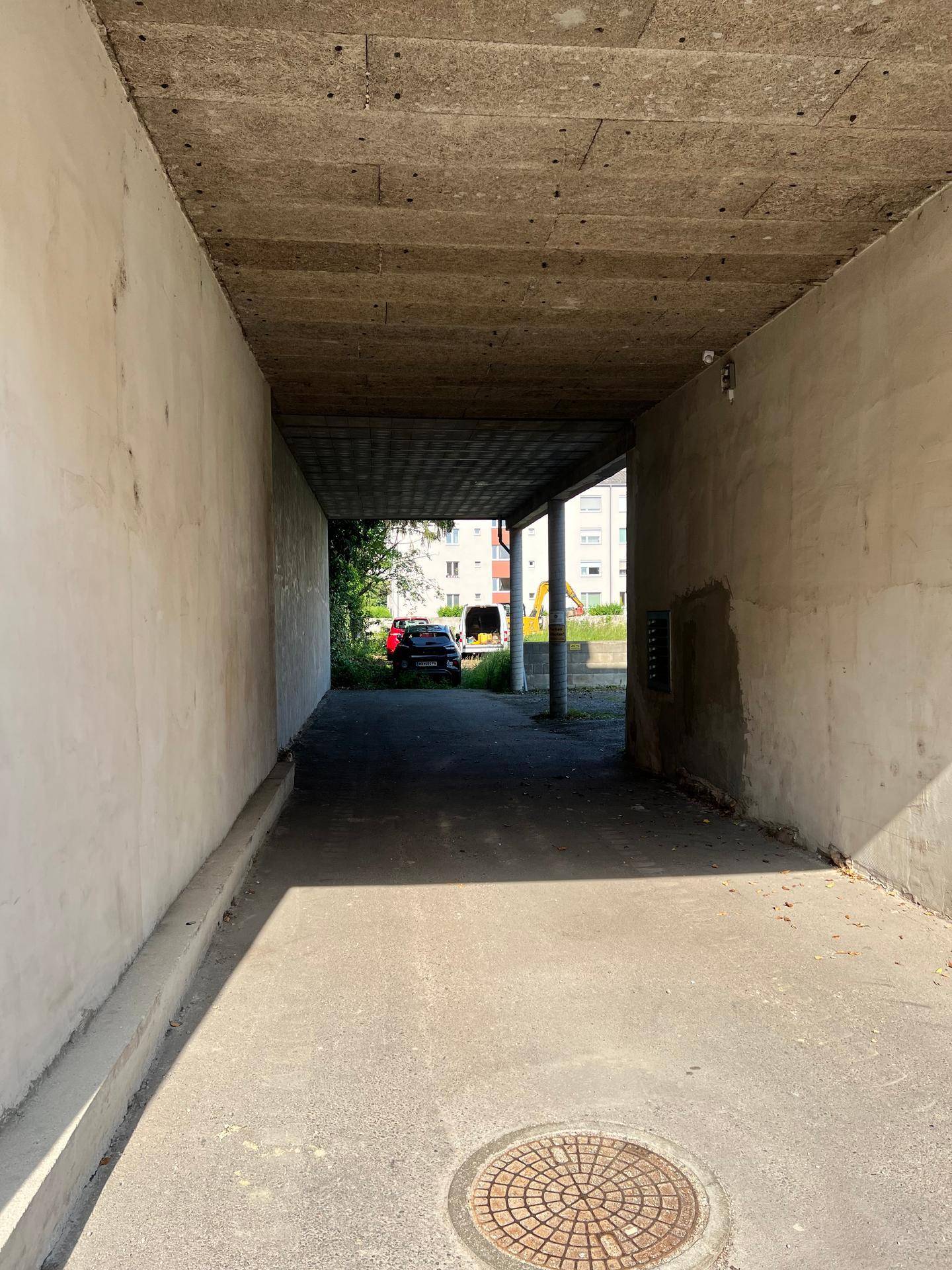 Betonierter Durchgang oder Unterführung mit Blick auf einen Parkplatz und Grünflächen.