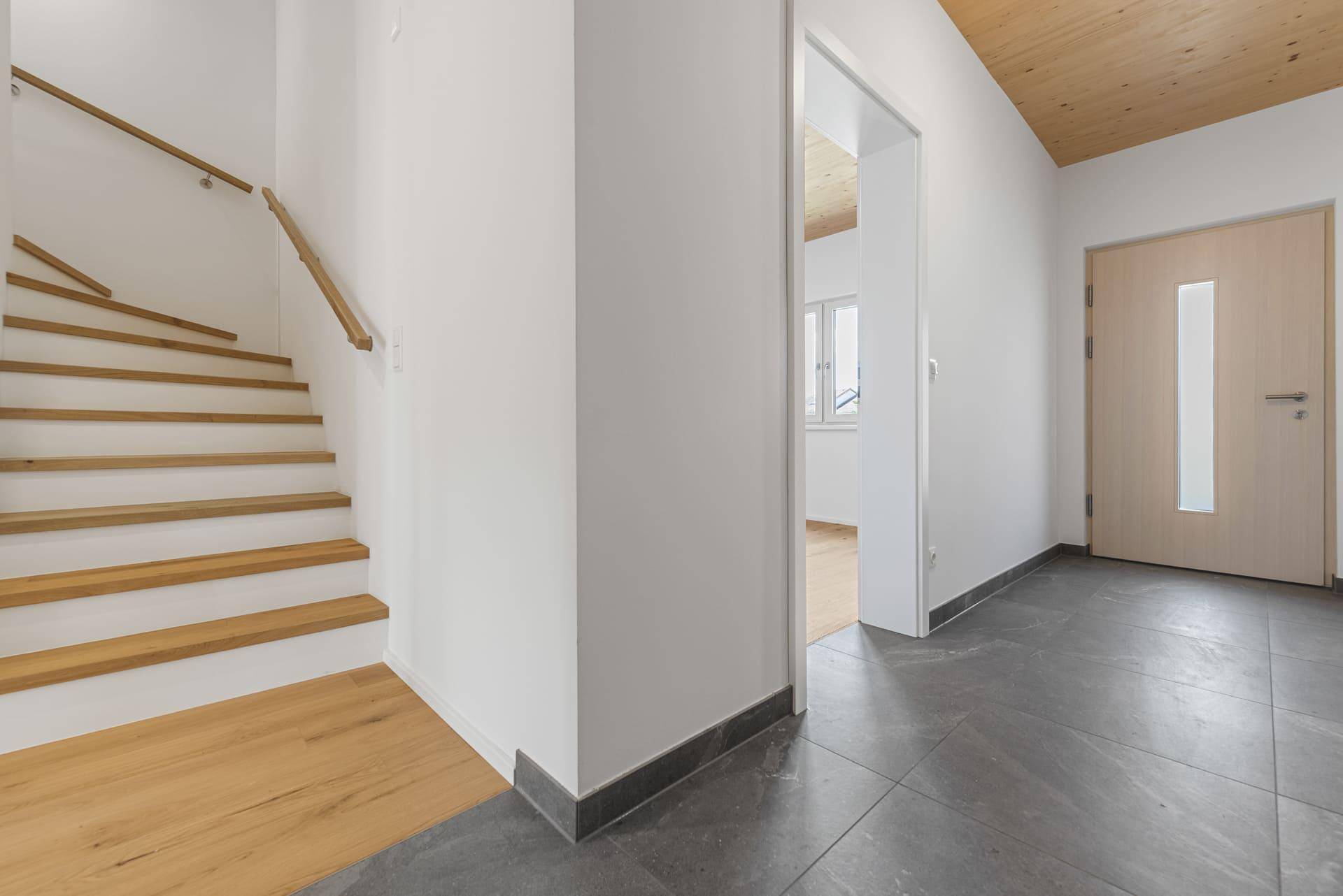 Eingangsbereich mit Treppe, grauen Fliesen, Holzboden und einer Holztür.