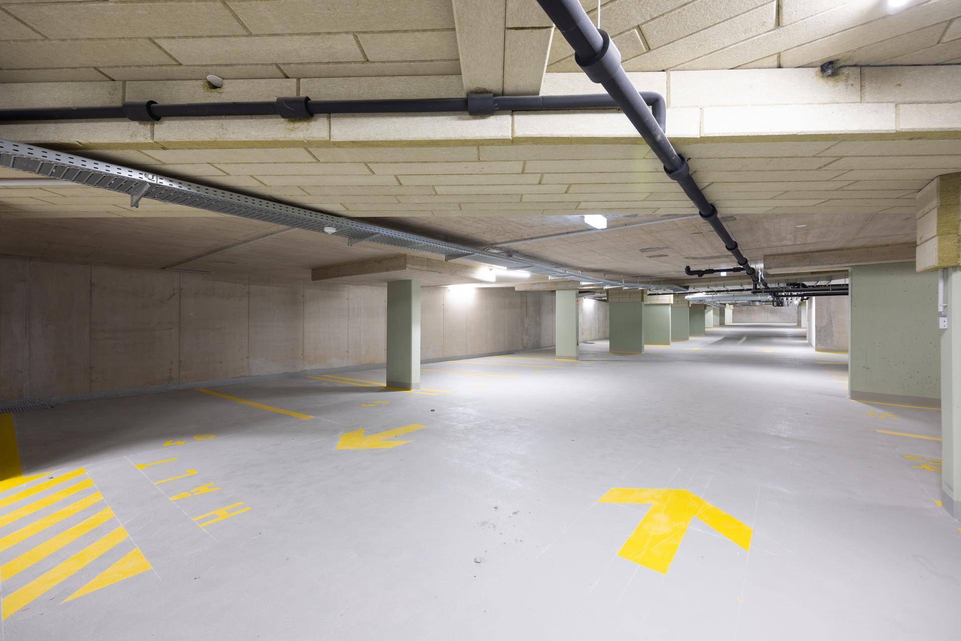 Weitläufige Tiefgarage mit gelben Markierungen auf dem Boden und sichtbaren Rohren an der Decke.
