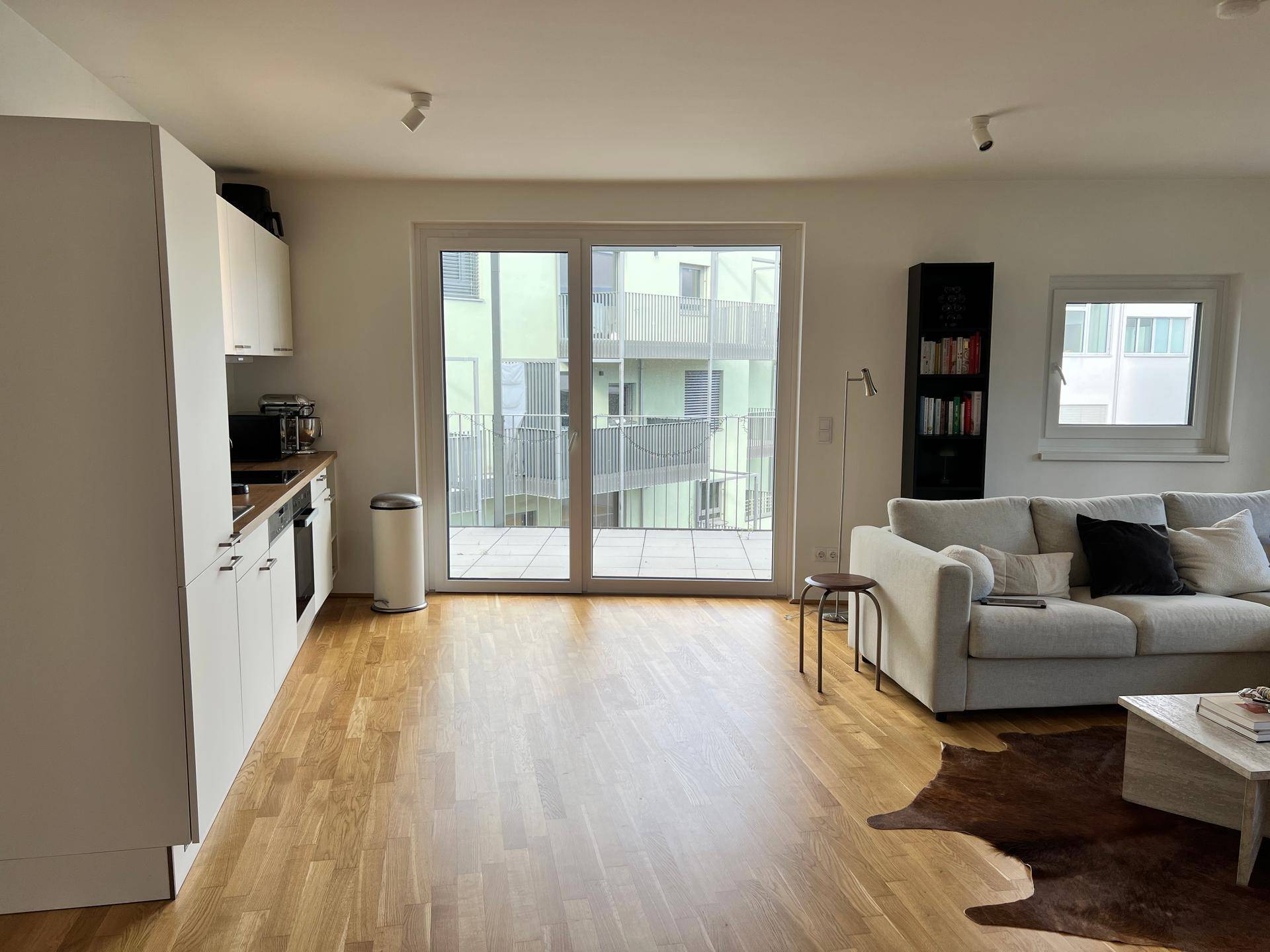 Offener Wohnbereich mit Holzboden, Sofa und Blick auf die moderne Küche und den Balkon.