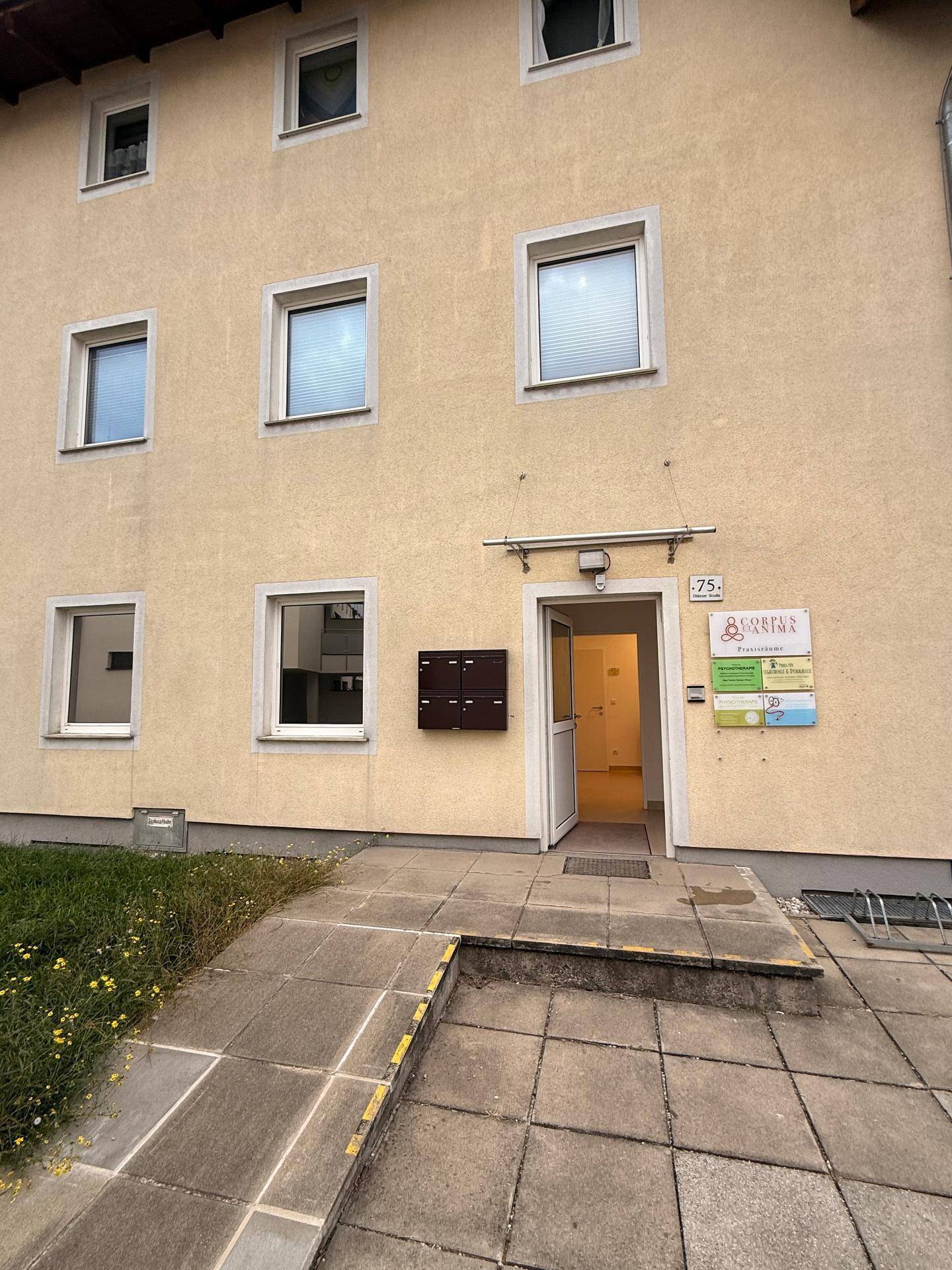 Gebäudefassade mit Eingangsbereich, Rampe für Barrierefreiheit und mehreren Fenstern.