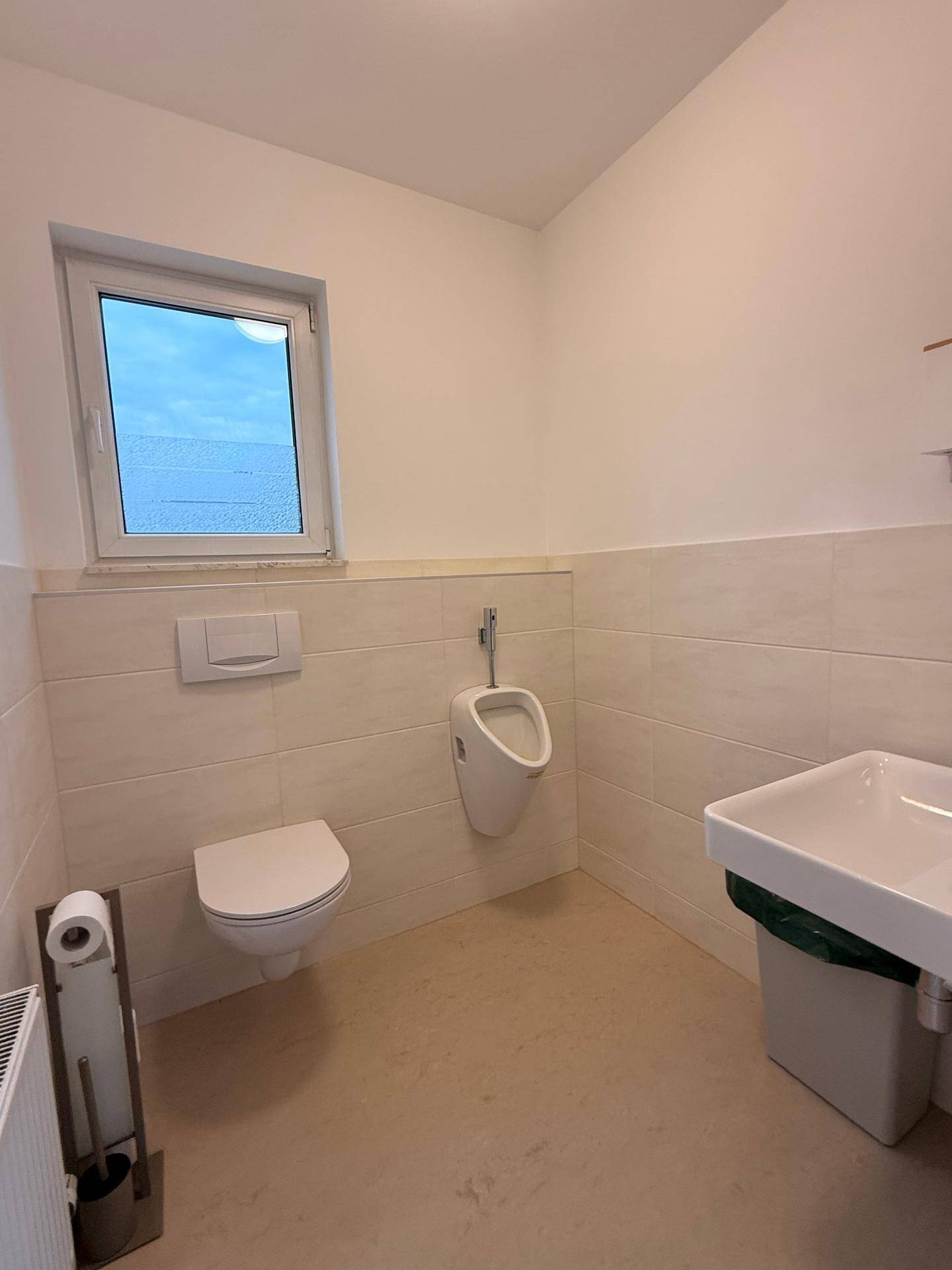 Sauberer Toilettenraum mit Fenster, Urinal, wandhängendem WC und hellem Fliesenboden.