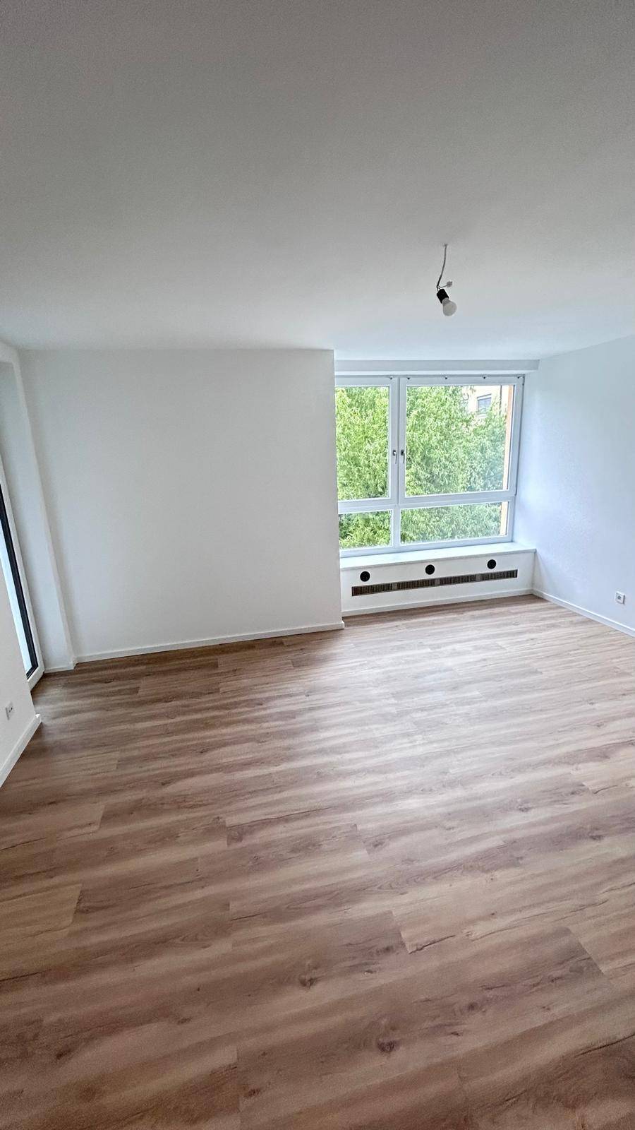 Helles Zimmer mit Laminatboden, weißen Wänden und einem Fenster mit Blick ins Grüne.