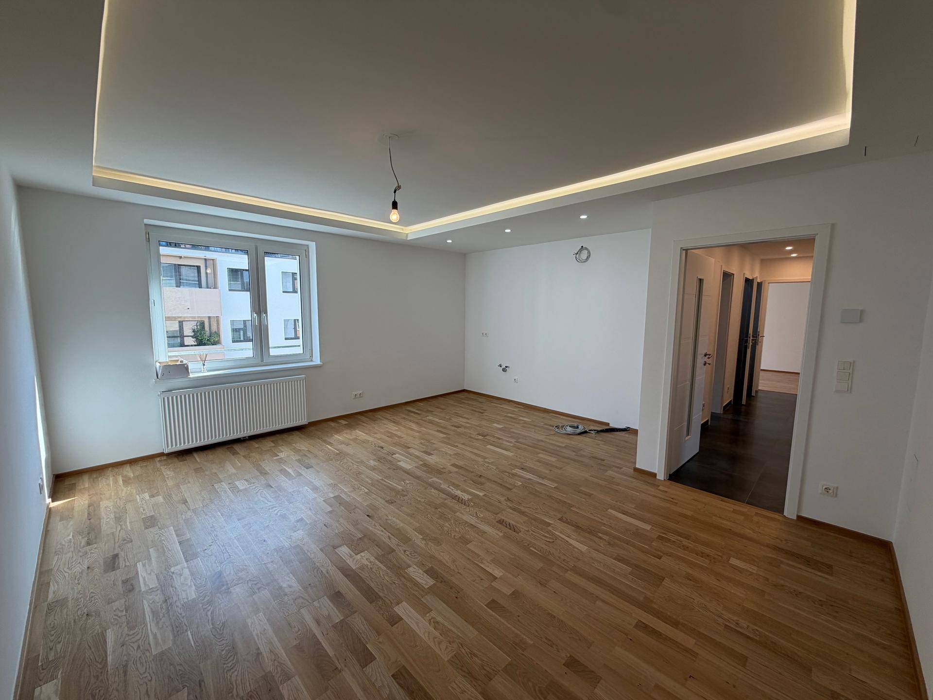 Geräumiges, leeres Wohnzimmer mit hellem Parkettboden, weißen Wänden und moderner Deckenbeleuchtung.
