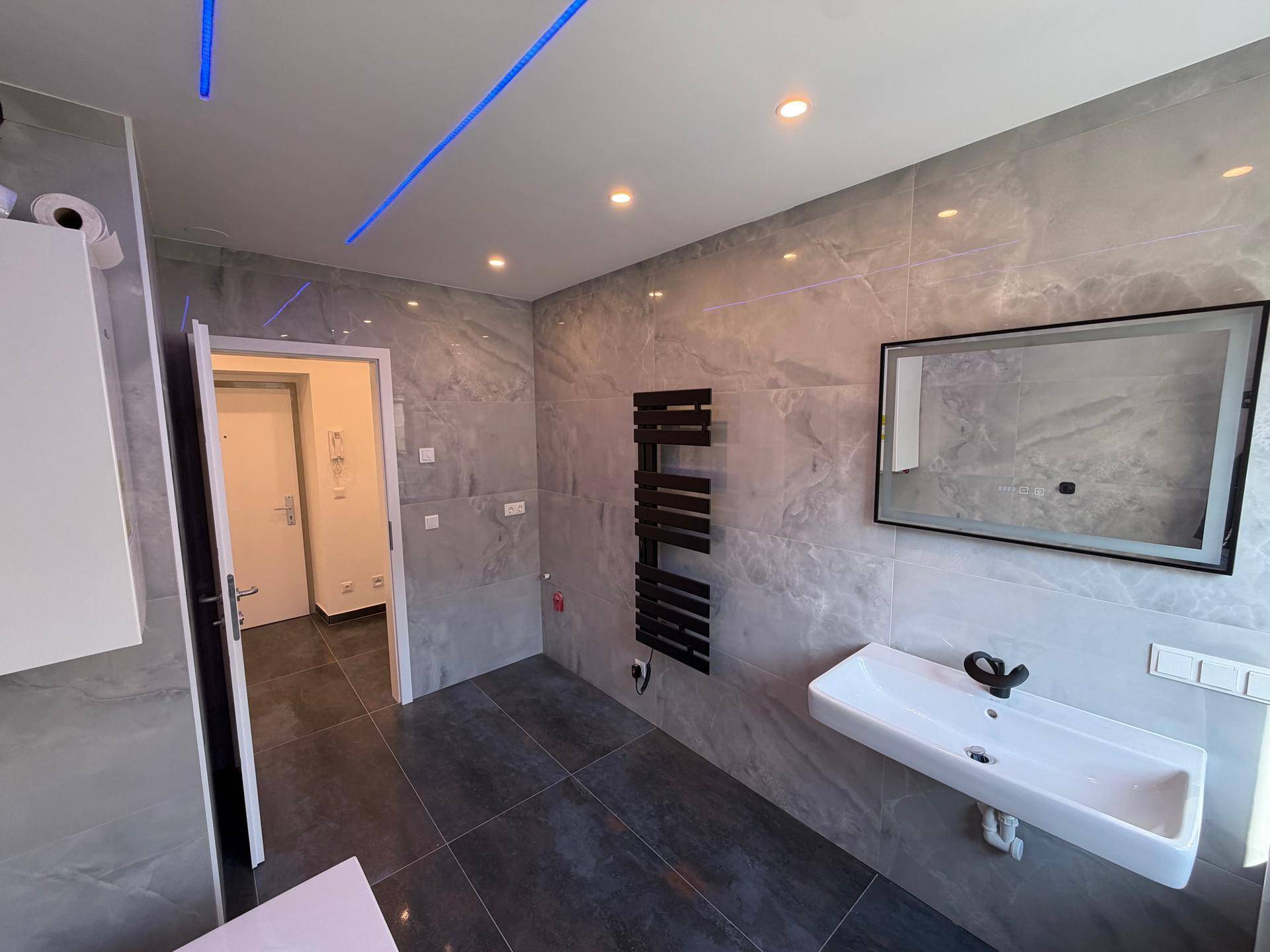 Helles Badezimmer mit modernen grauen Fliesen, Waschbecken, Spiegel und beheiztem Handtuchhalter.