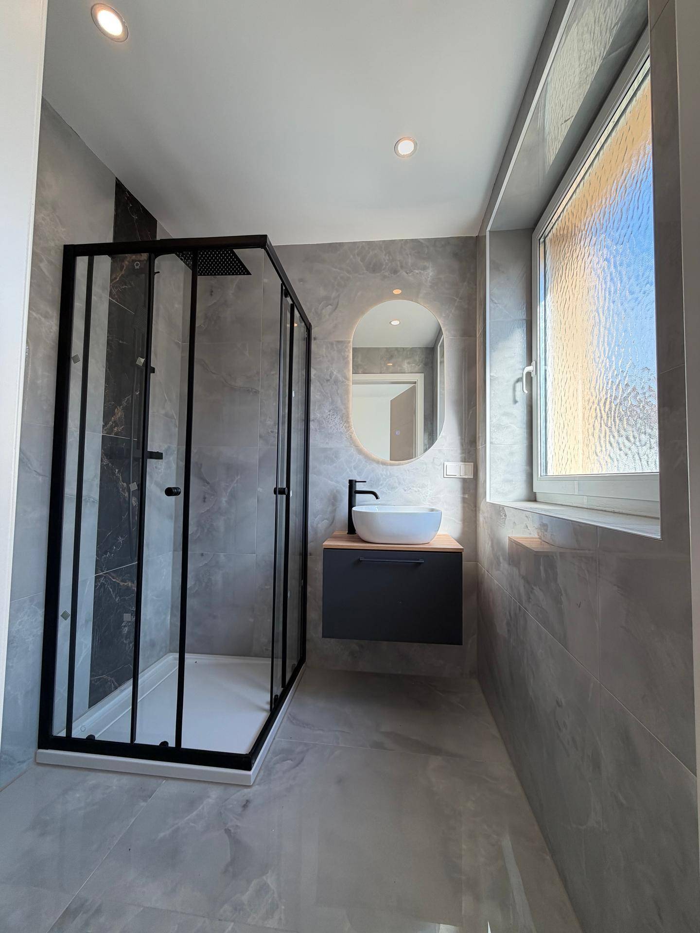 Modernes Badezimmer mit Duschkabine, Waschbecken auf Waschtisch und Fenster, in hellen Grautönen gehalten.