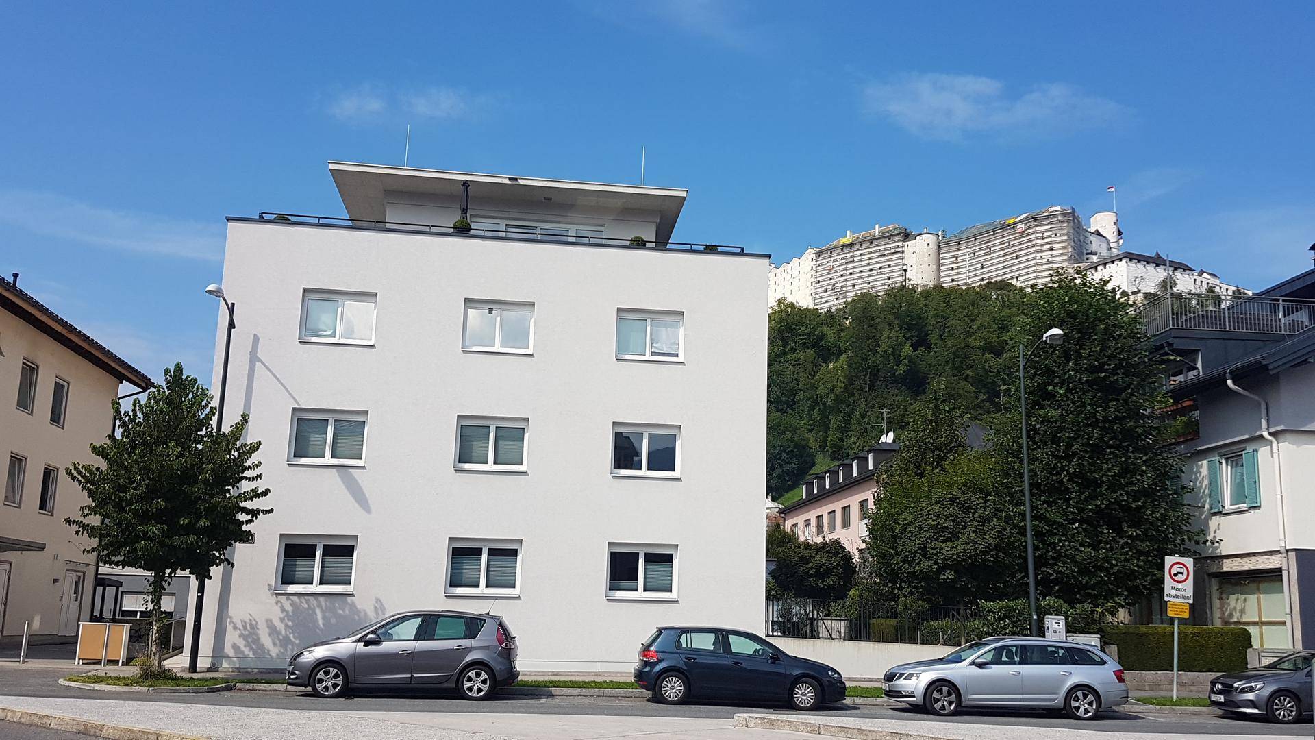Modernes Mehrfamilienhaus mit Flachdach und Blick auf eine bewaldete Anhöhe.