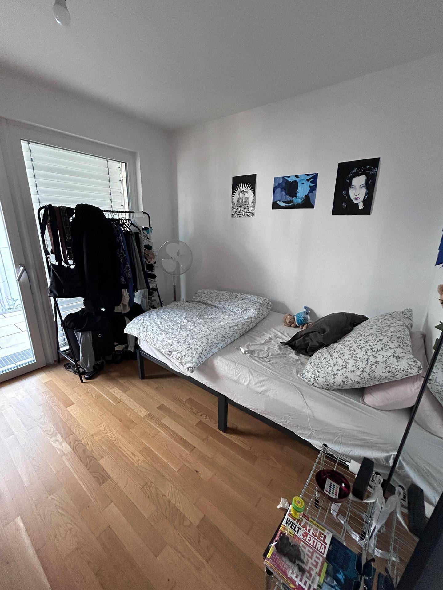 Schlafzimmer mit Holzboden, einem Bett und einem Kleiderständer neben der Balkontür.