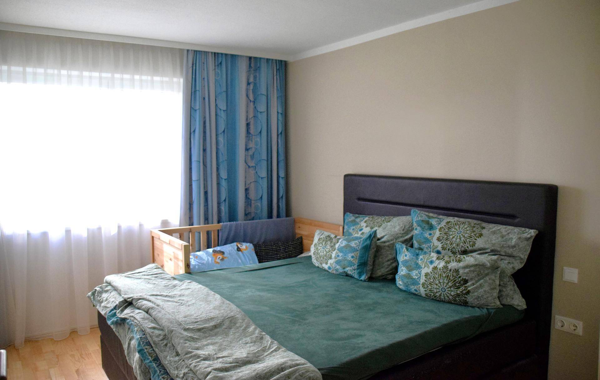 Helles Schlafzimmer mit großem Bett, blauen Vorhängen und einem Babybett am Fenster.