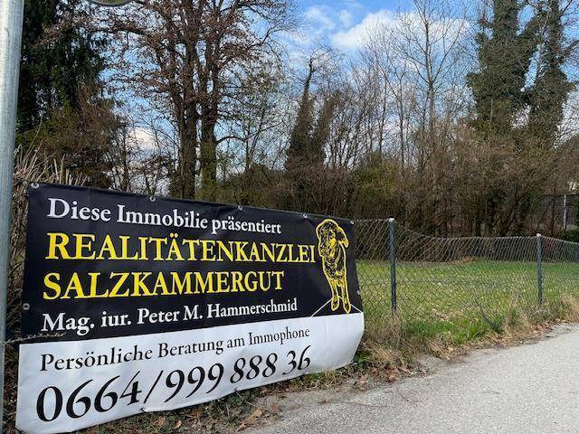 Ein großes Maklerschild am Straßenrand vor einem Zaun und einer grünen Fläche mit Bäumen.