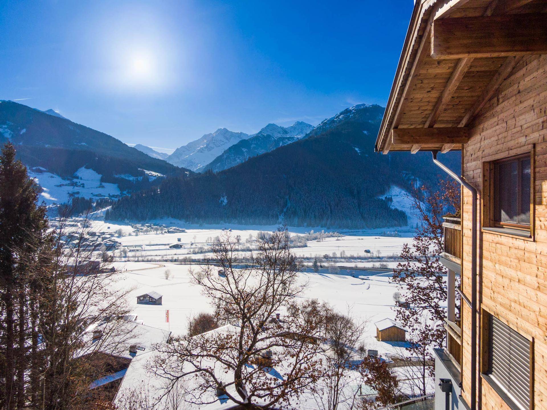 Sonniger Blick auf die verschneite Berglandschaft und das Tal von der Seite eines Holzchalets.