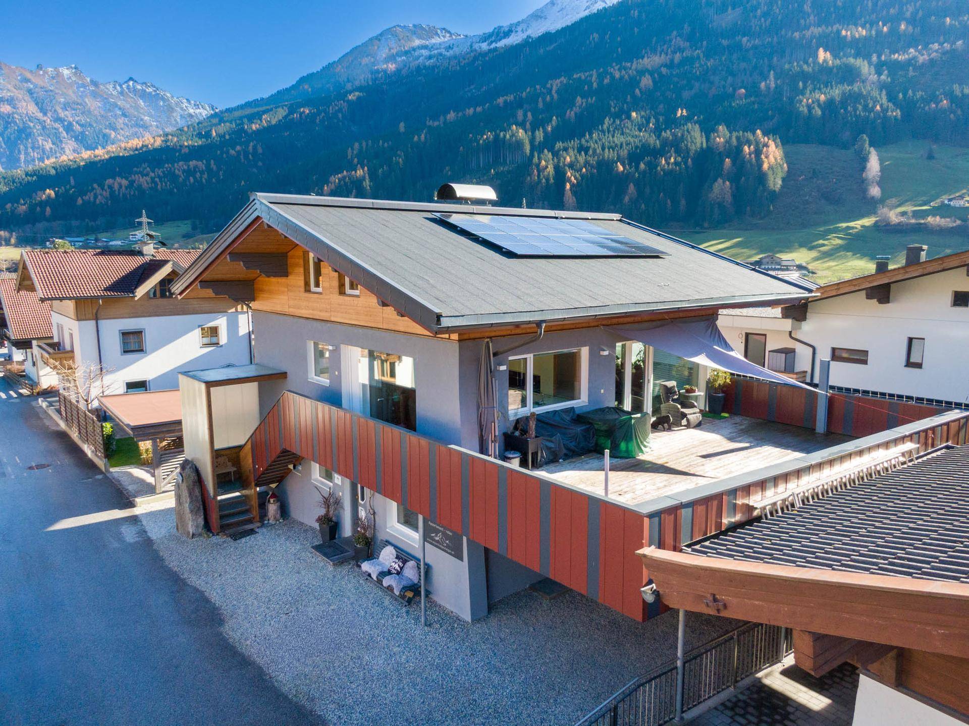 Gebäudefassade mit großer Terrasse und Solarpaneelen auf dem Dach in sonniger Berglandschaft.