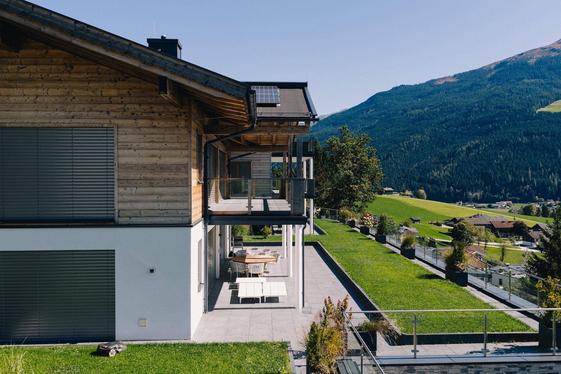 Seitlicher Blick auf ein modernes Holzhaus mit großer Terrasse, Garten und Bergblick.