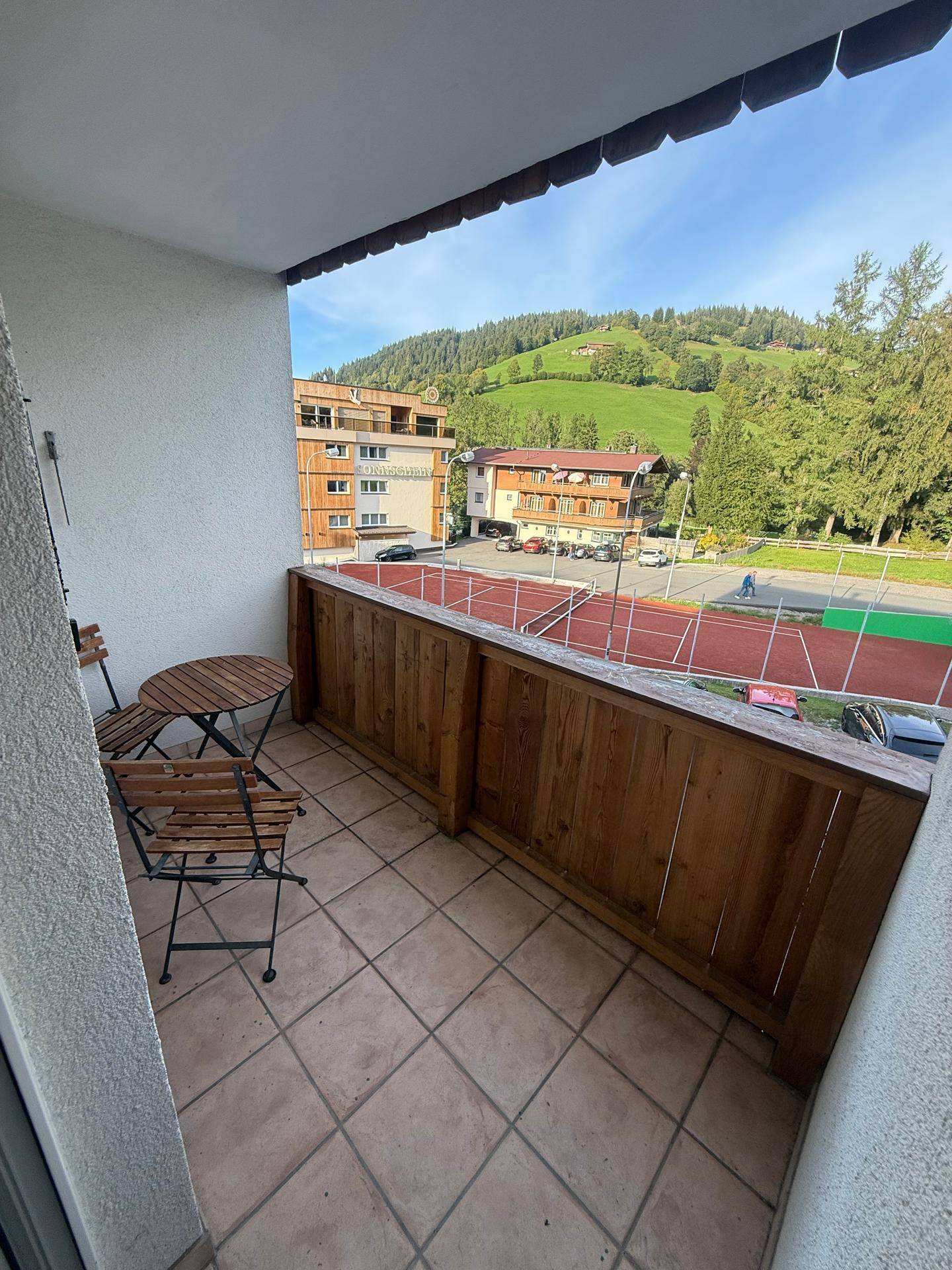 Balkon mit Holztisch und Stühlen, Blick auf Tennisplatz und Umgebung.