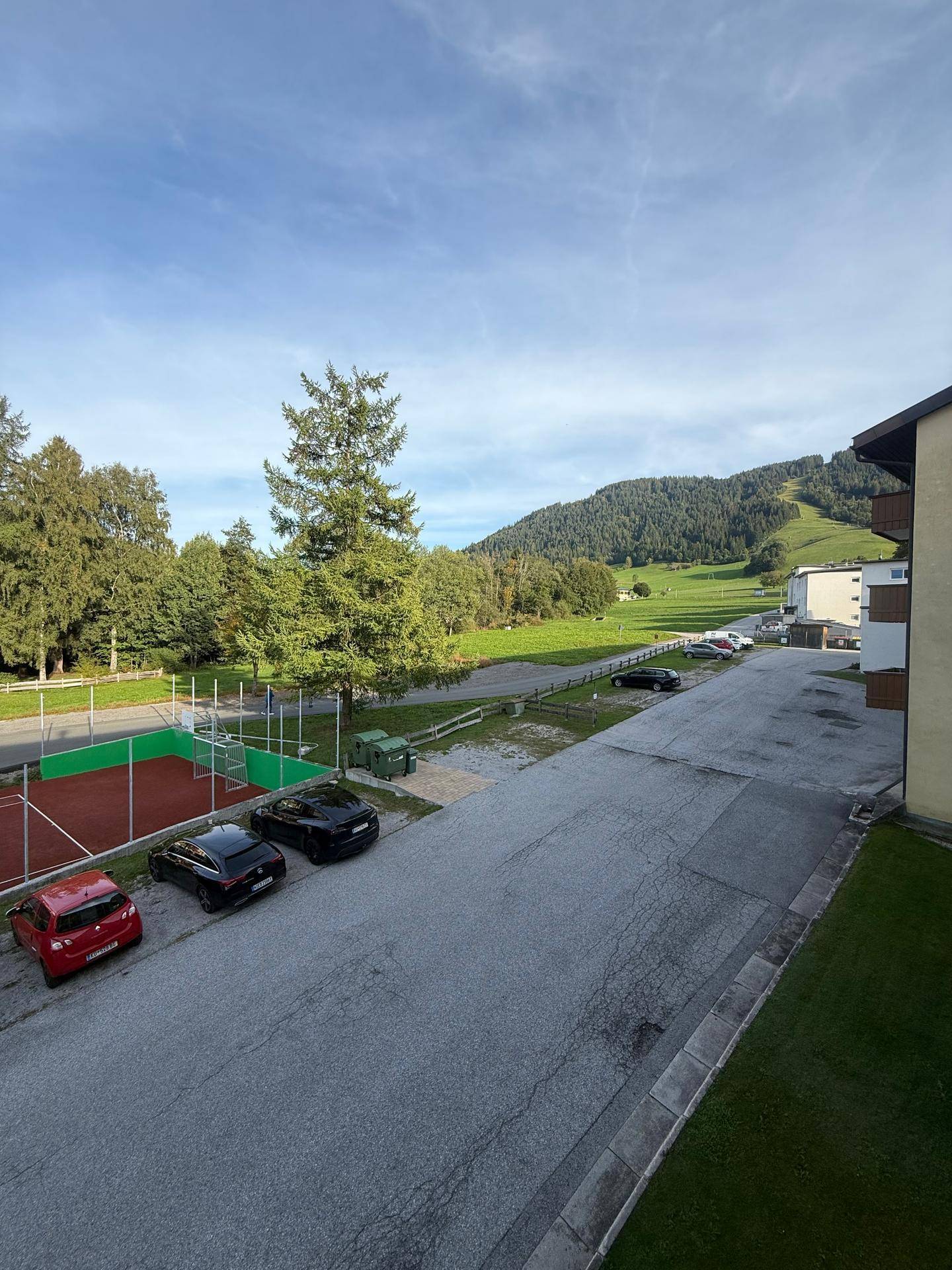 Außenansicht mit Tennisplatz, Parkplätzen und grüner Berglandschaft im Hintergrund.