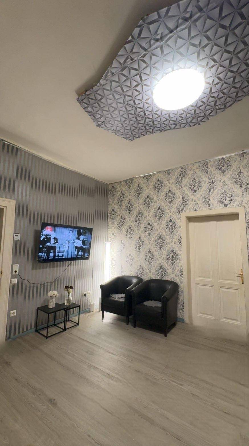 Wohnzimmer mit Wandfernseher, zwei schwarzen Sesseln und gemusterter Tapete, beleuchtet von einer Deckenlampe.