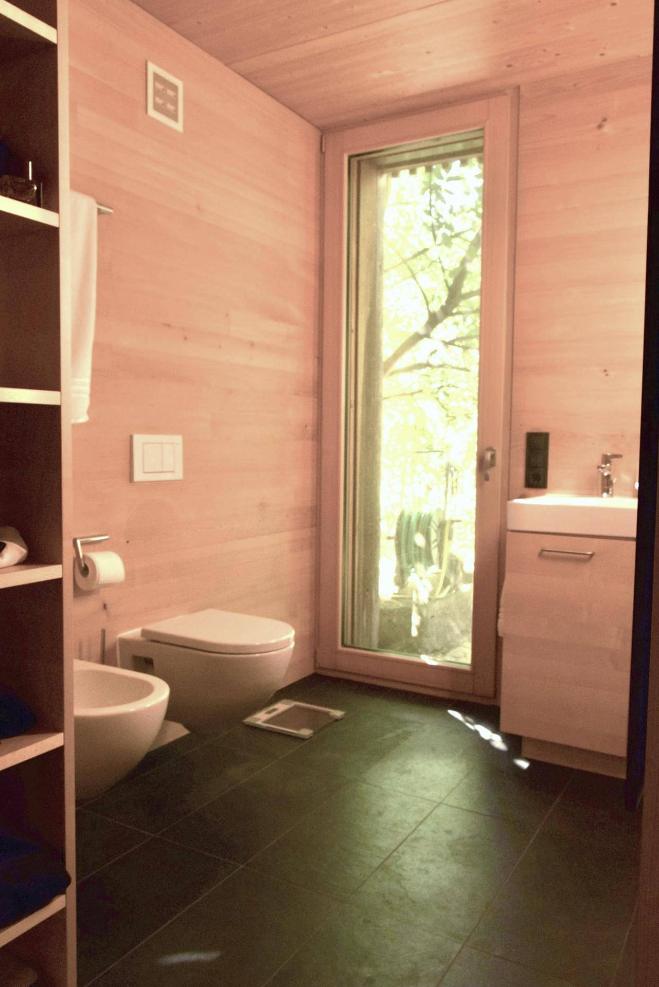 Modernes Badezimmer mit Holzwänden, dunklen Fliesen, WC, Bidet und Fenster.