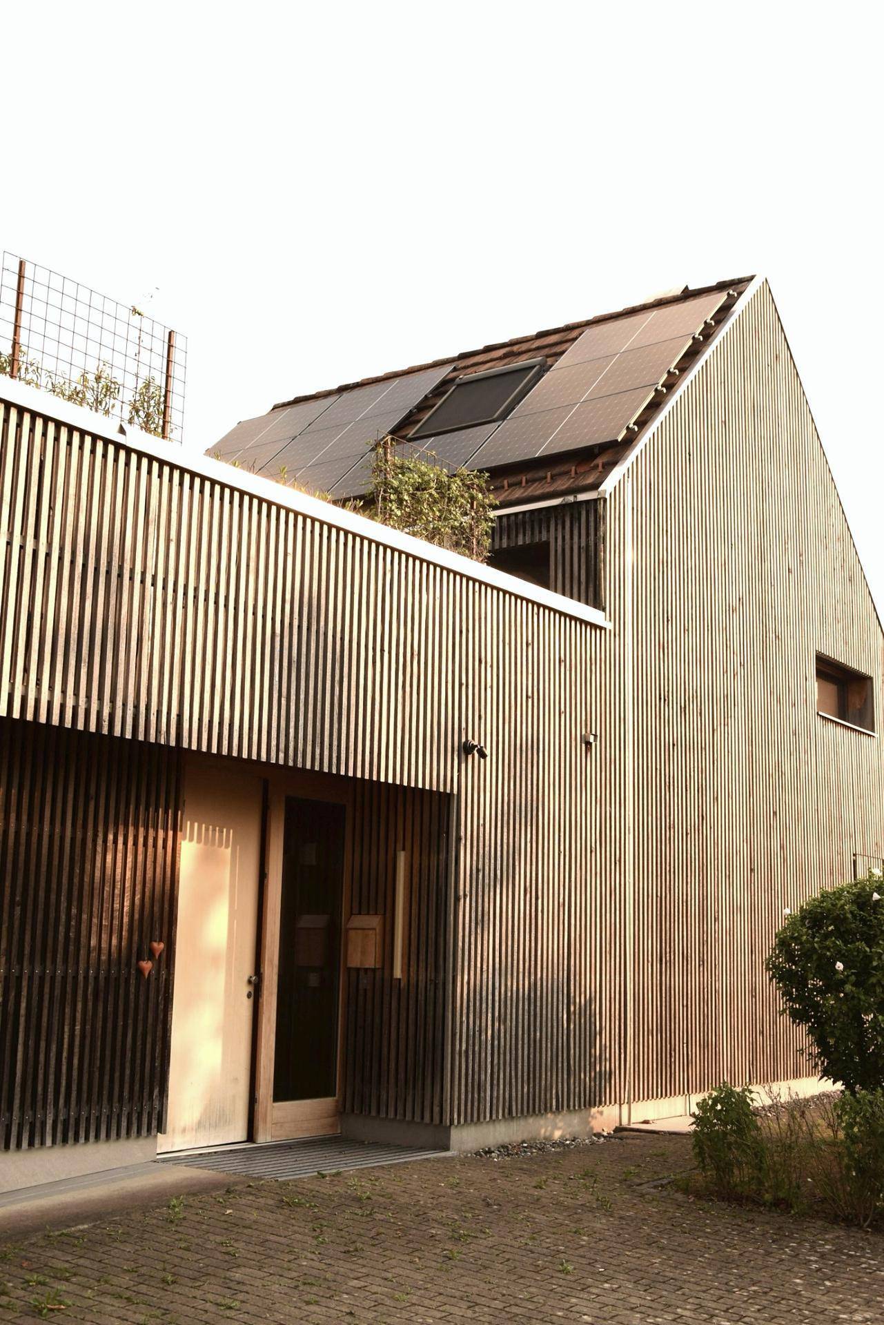 Fassade eines modernen Holzhauses mit vertikalen Lamellen und Solarpaneelen auf dem Dach.