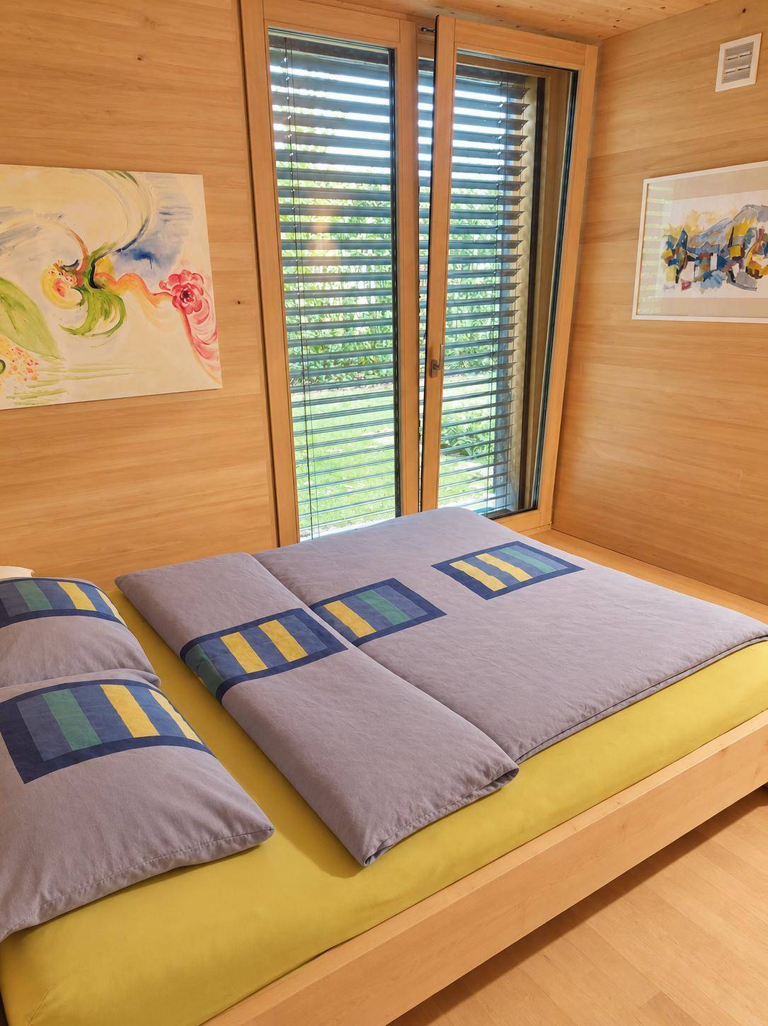 Helles Schlafzimmer mit Holzwänden, farbiger Bettwäsche und Blick ins Grüne.