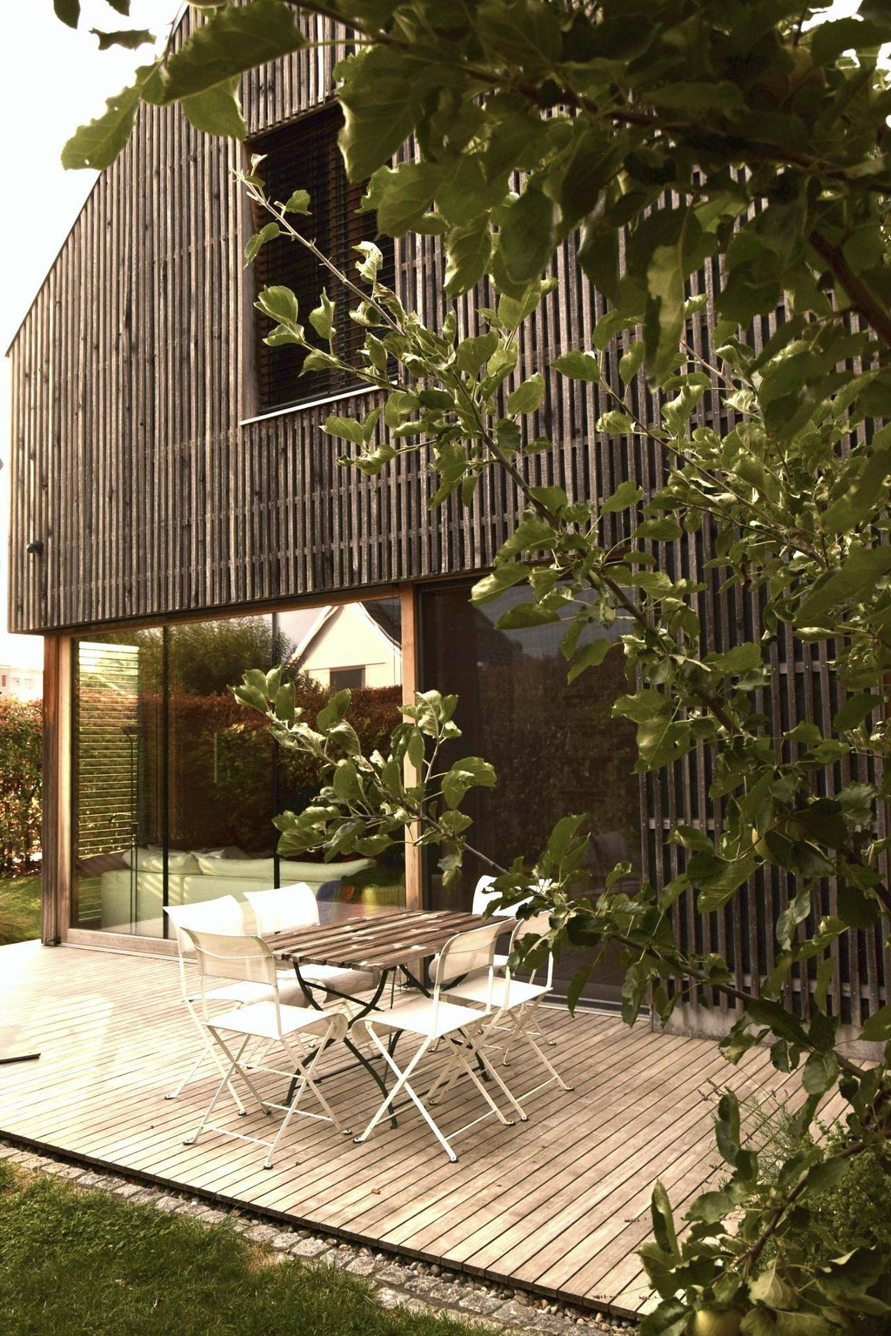 Holzterrasse mit Gartenmöbeln neben einem modernen Holzhaus, umgeben von Bäumen.