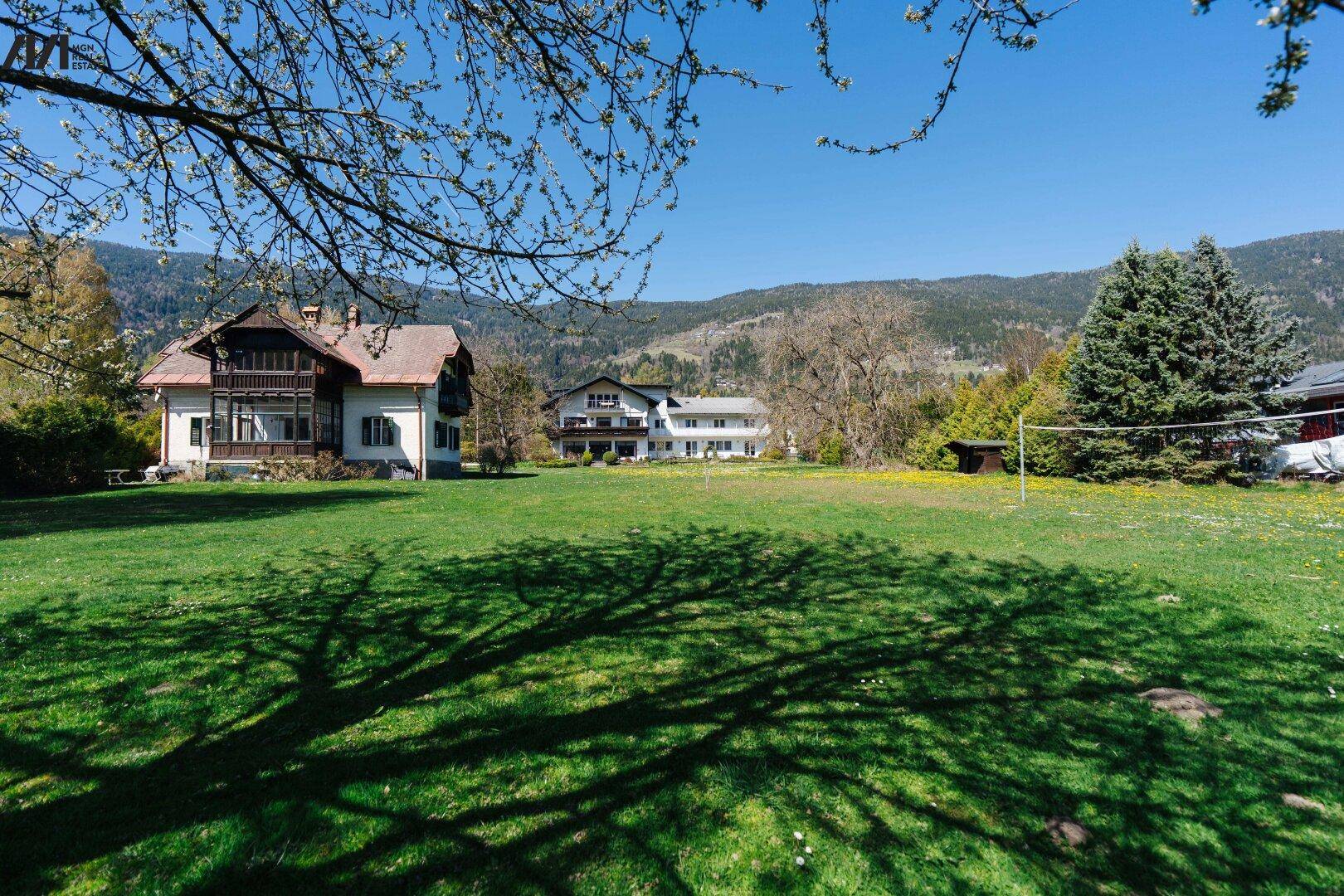 Großer grüner Garten mit altem Baumbestand, einem traditionellen Haus und Volleyballnetz vor Bergkulisse.