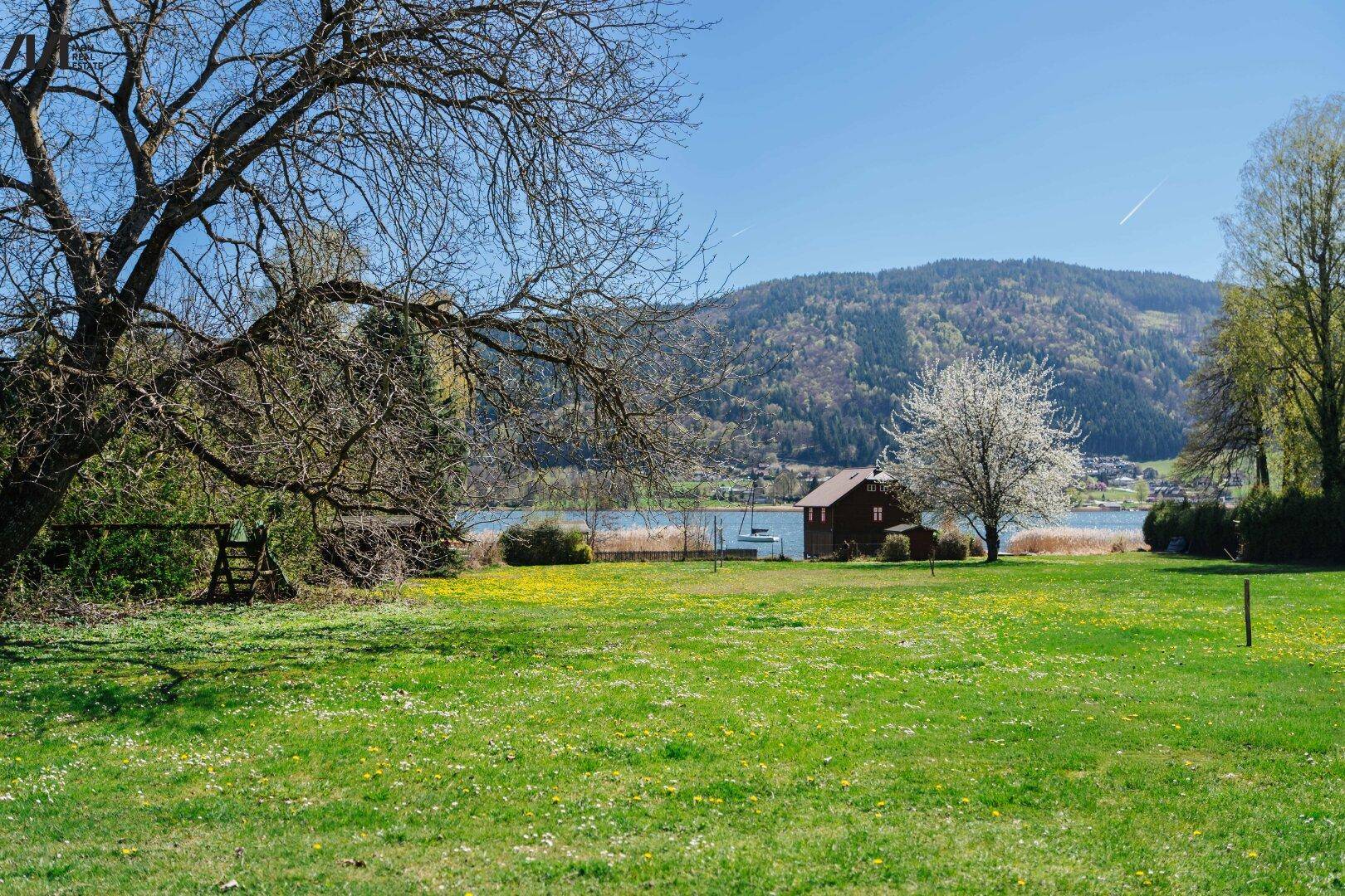 Weitläufige Wiese mit blühenden Bäumen, einem Bootshaus am Seeufer und sanften Hügeln im Hintergrund.